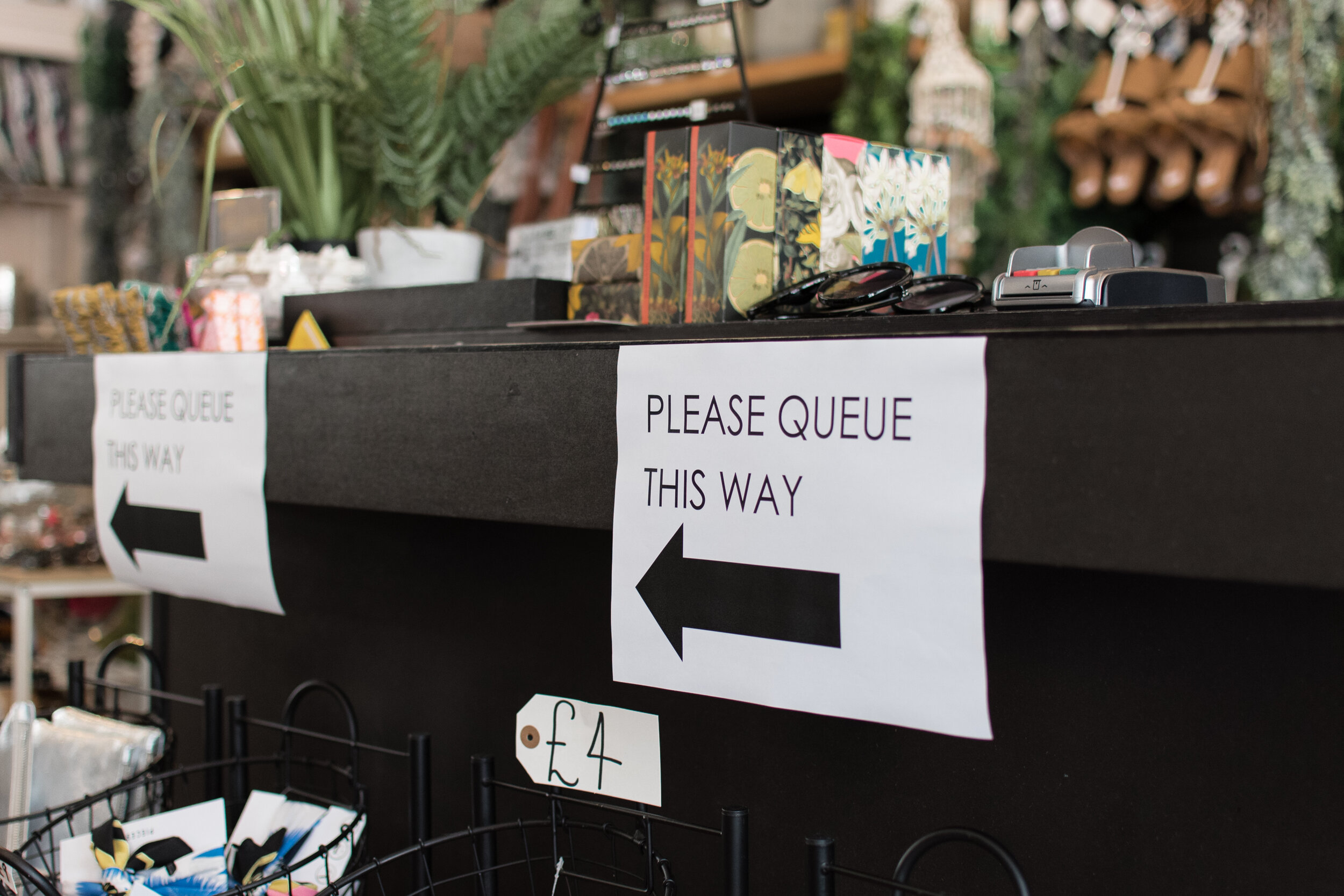 Counter with 'please queue this way' sign and arrow, cash register, sunglasses, and decorative items in a store.