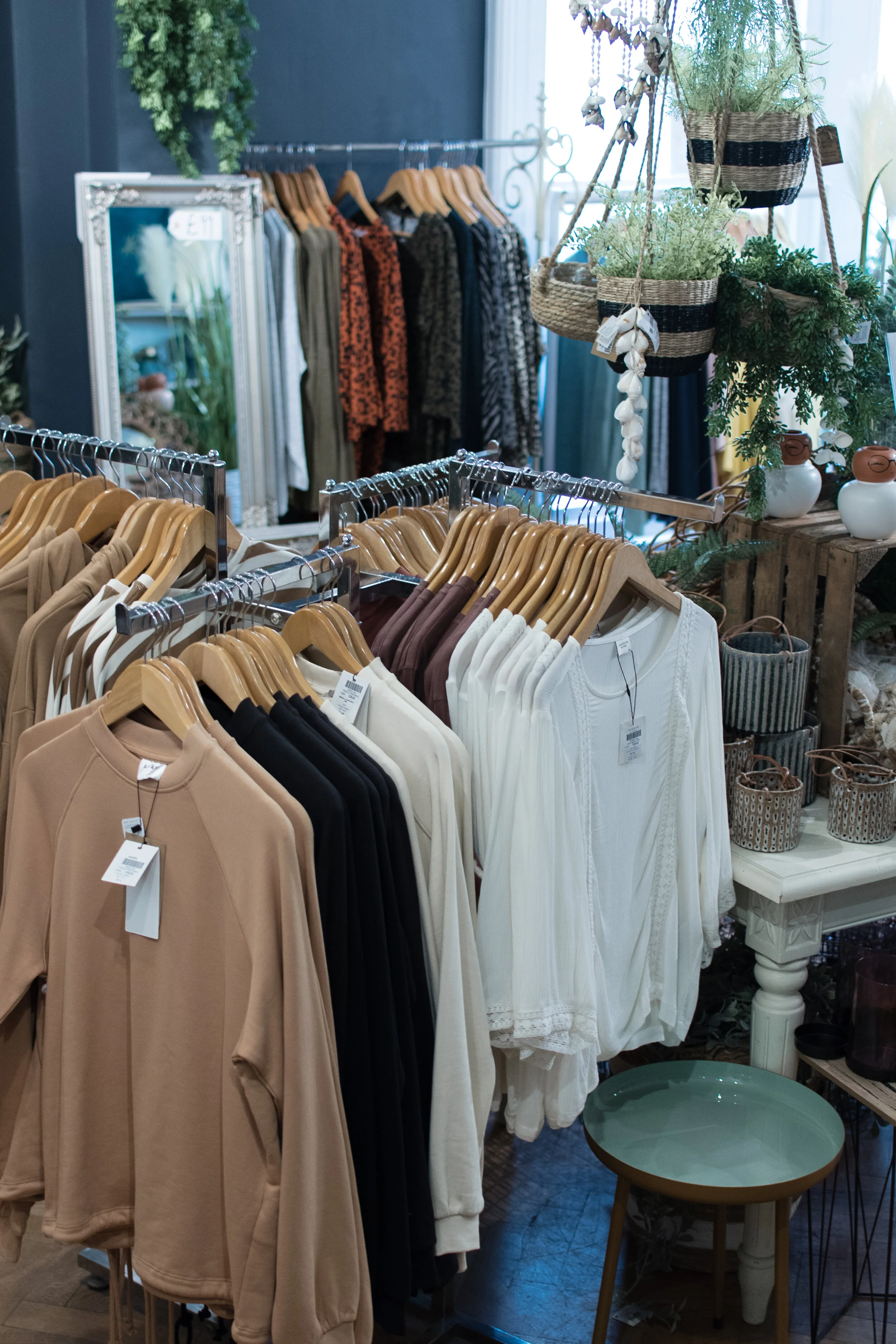 Clothing store interior with racks of shirts and sweaters on hangers, surrounded by decorative plants and decor items.