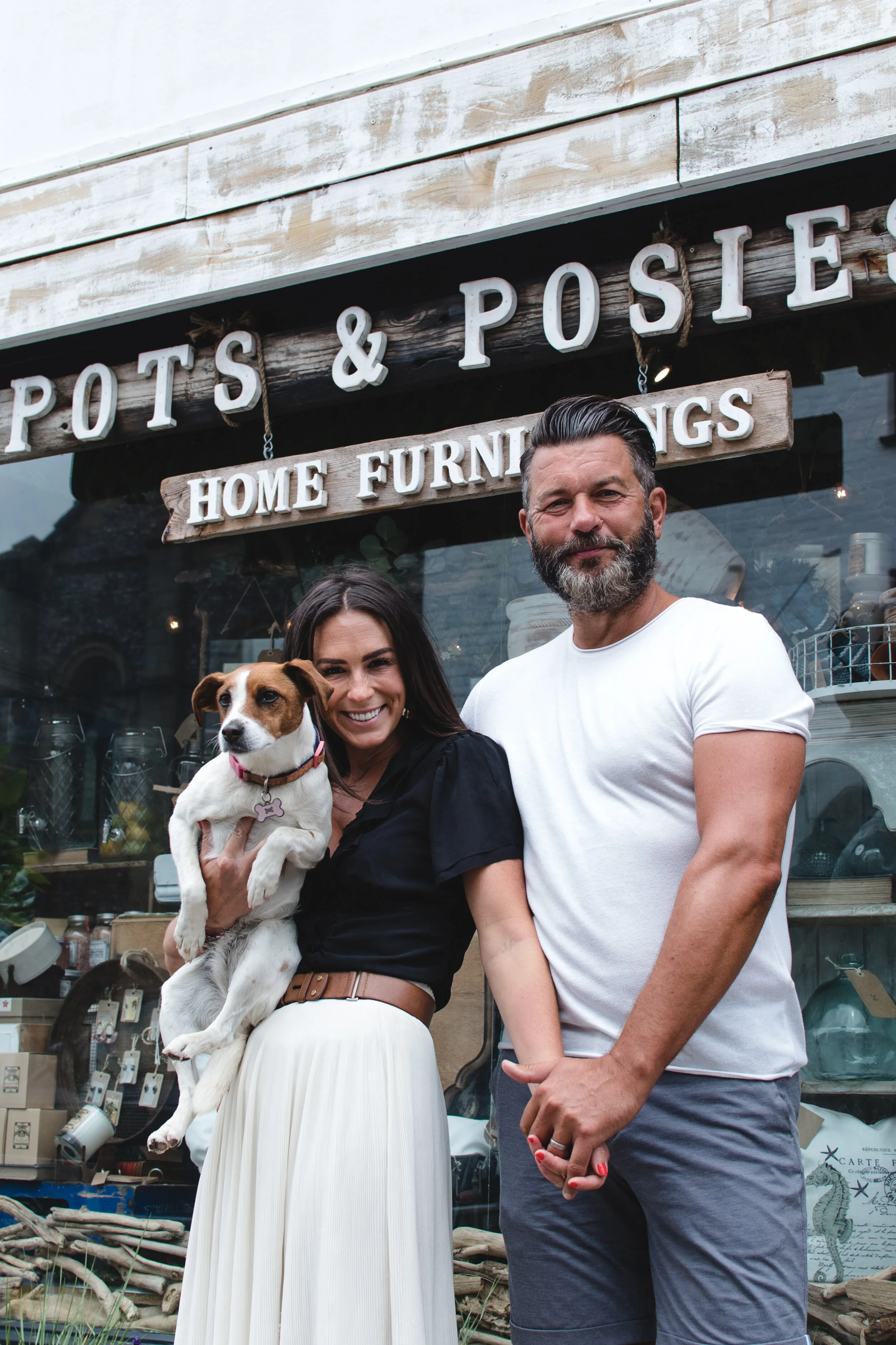 Two people standing in front of "Pots & Posies Home Furnishings" store, holding a small dog.