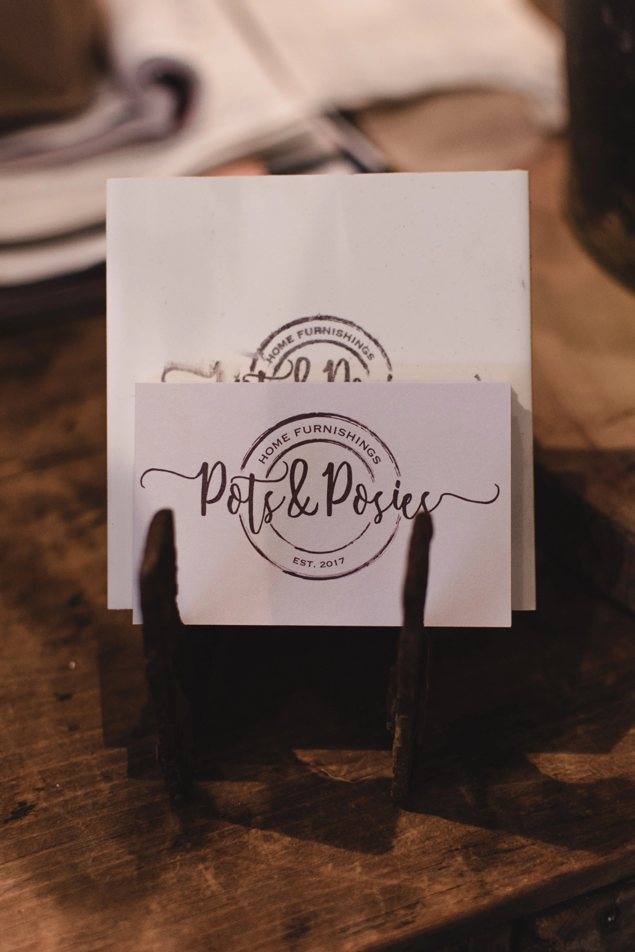 Business cards for "Pots & Posies" home furnishings, established 2017, displayed on a wooden surface.
