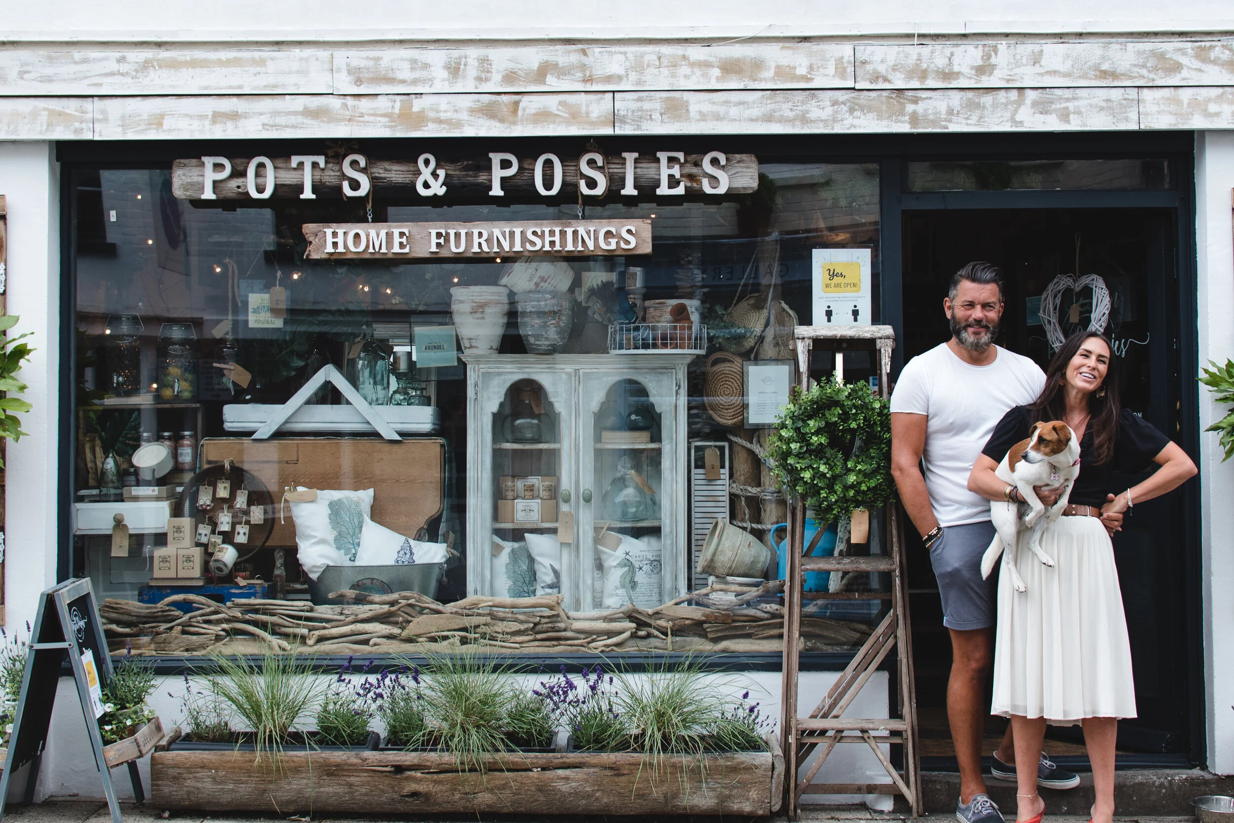 Exterior of 'Pots & Posies' home furnishings shop with two people standing in front, one holding a dog, surrounded by various home decor items and plants.