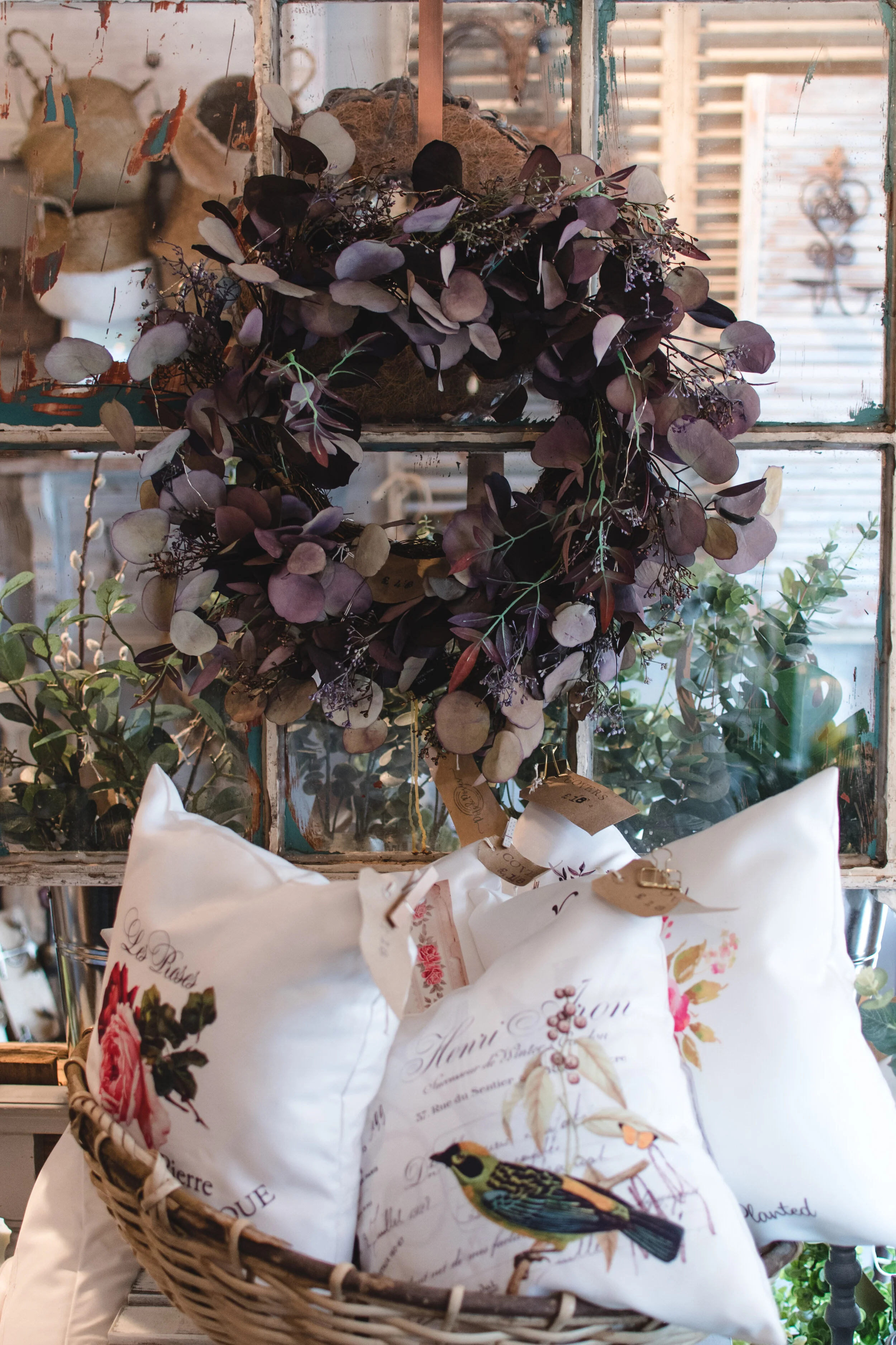 Rustic display with a purple wreath, botanical pillows, and vintage décor in a wooden basket on a distressed window background.