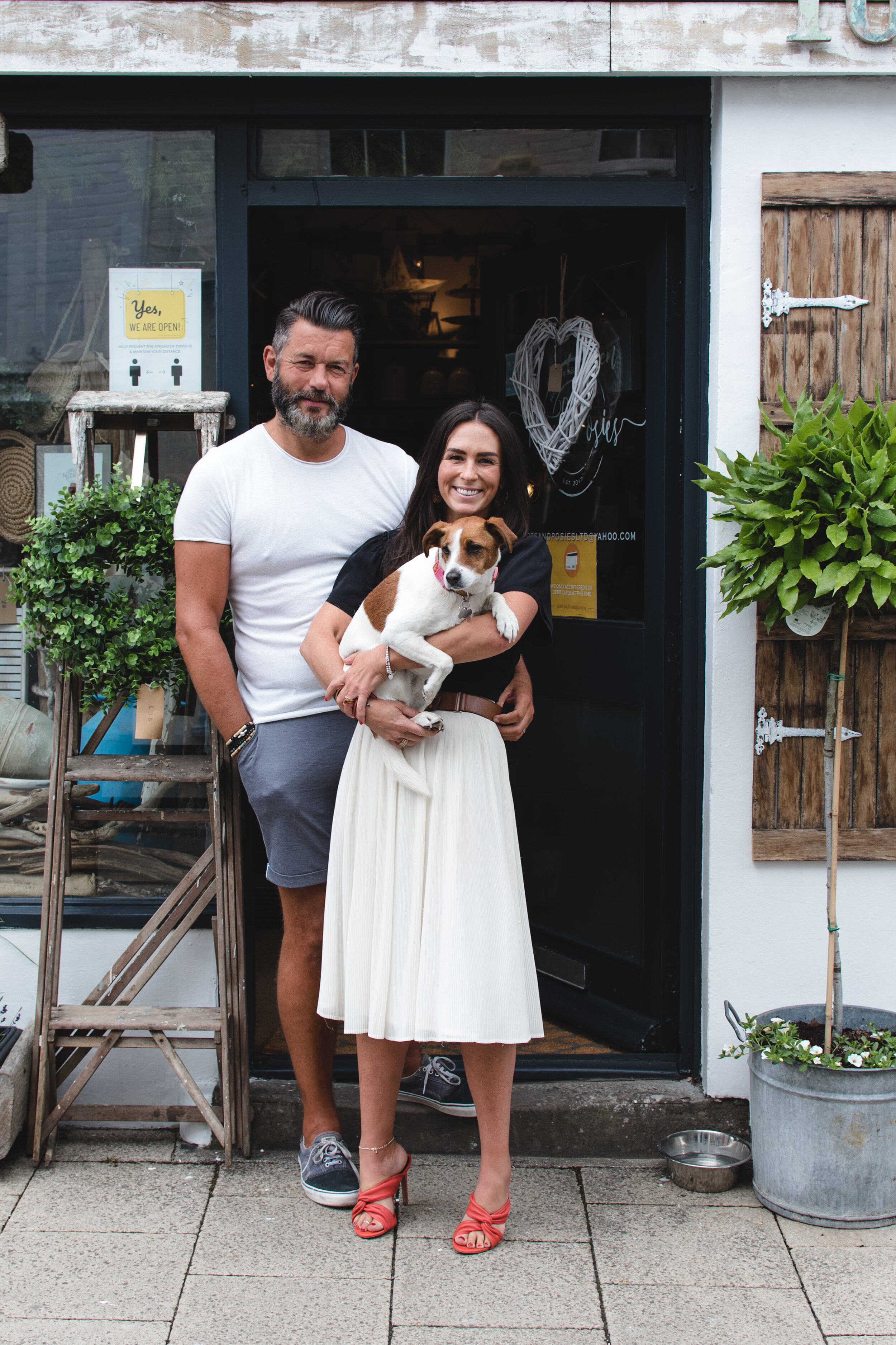 Couple standing outside a shop, woman holding a small dog, surrounded by plants.