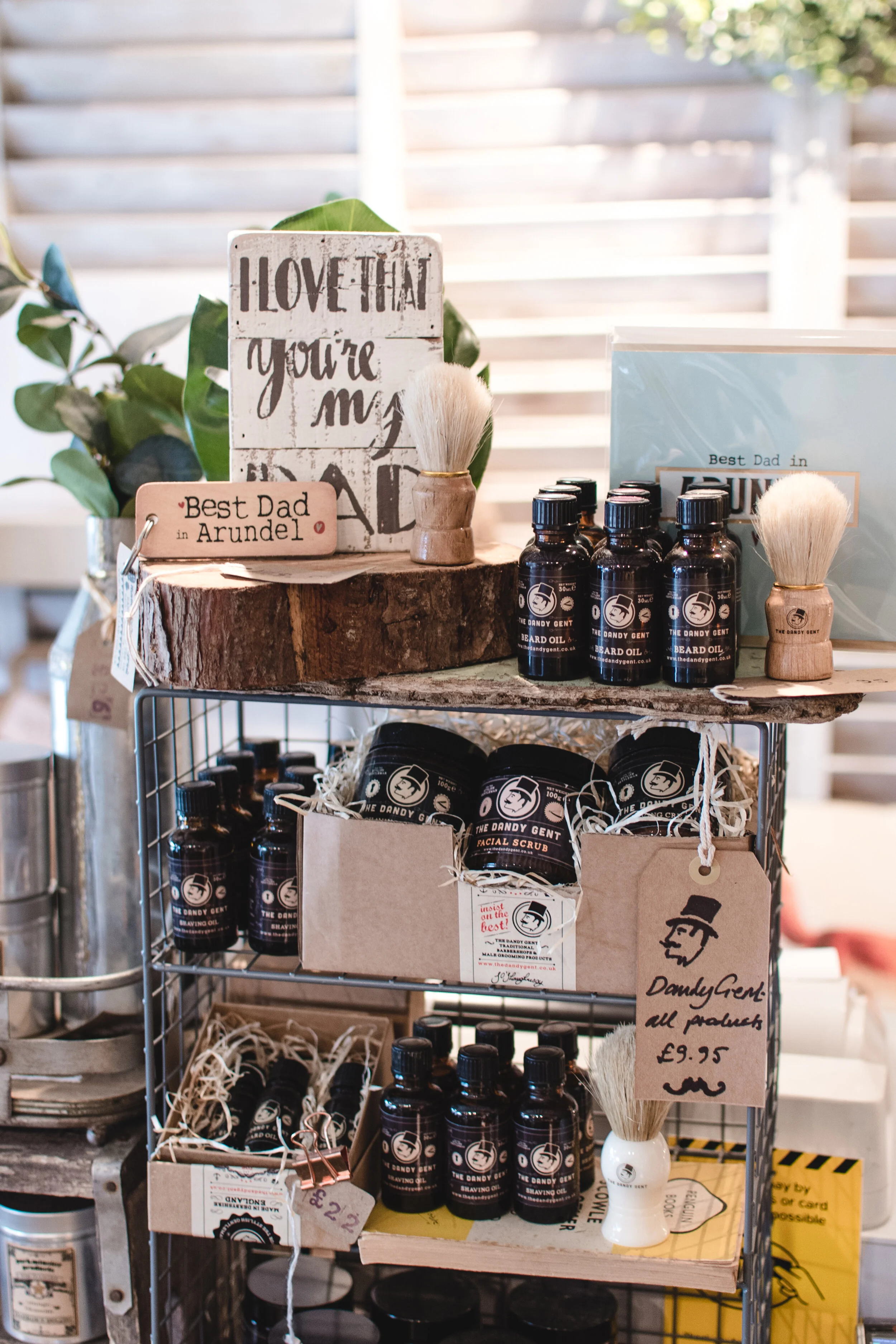 Display shelf with beard care products including beard oil and facial scrub from The Dandy Gent, shaving brushes, and decorative signs with phrases like 'Best Dad in Arundel.'