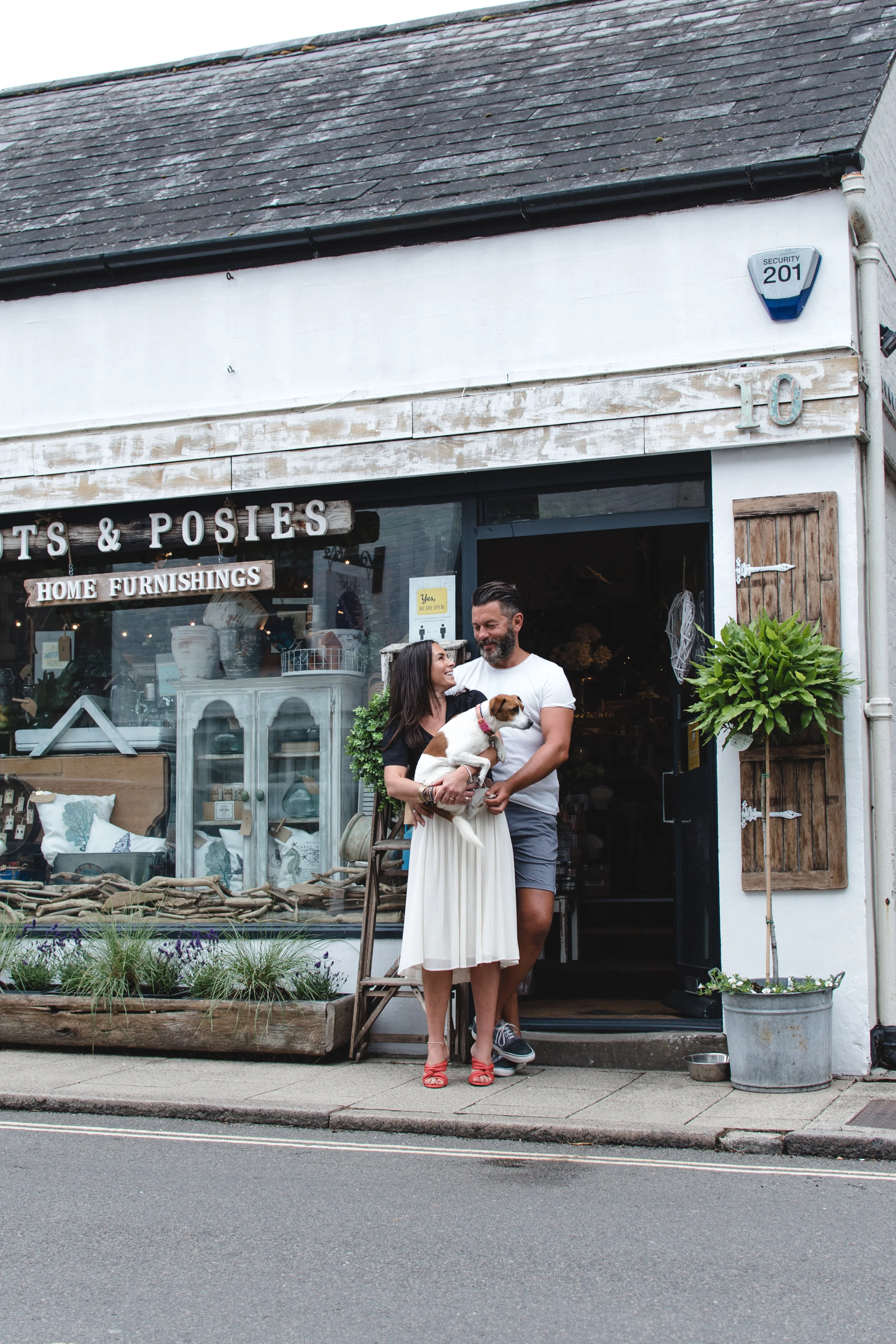 A couple standing in front of a home furnishings store, holding a dog, with a rustic storefront display.