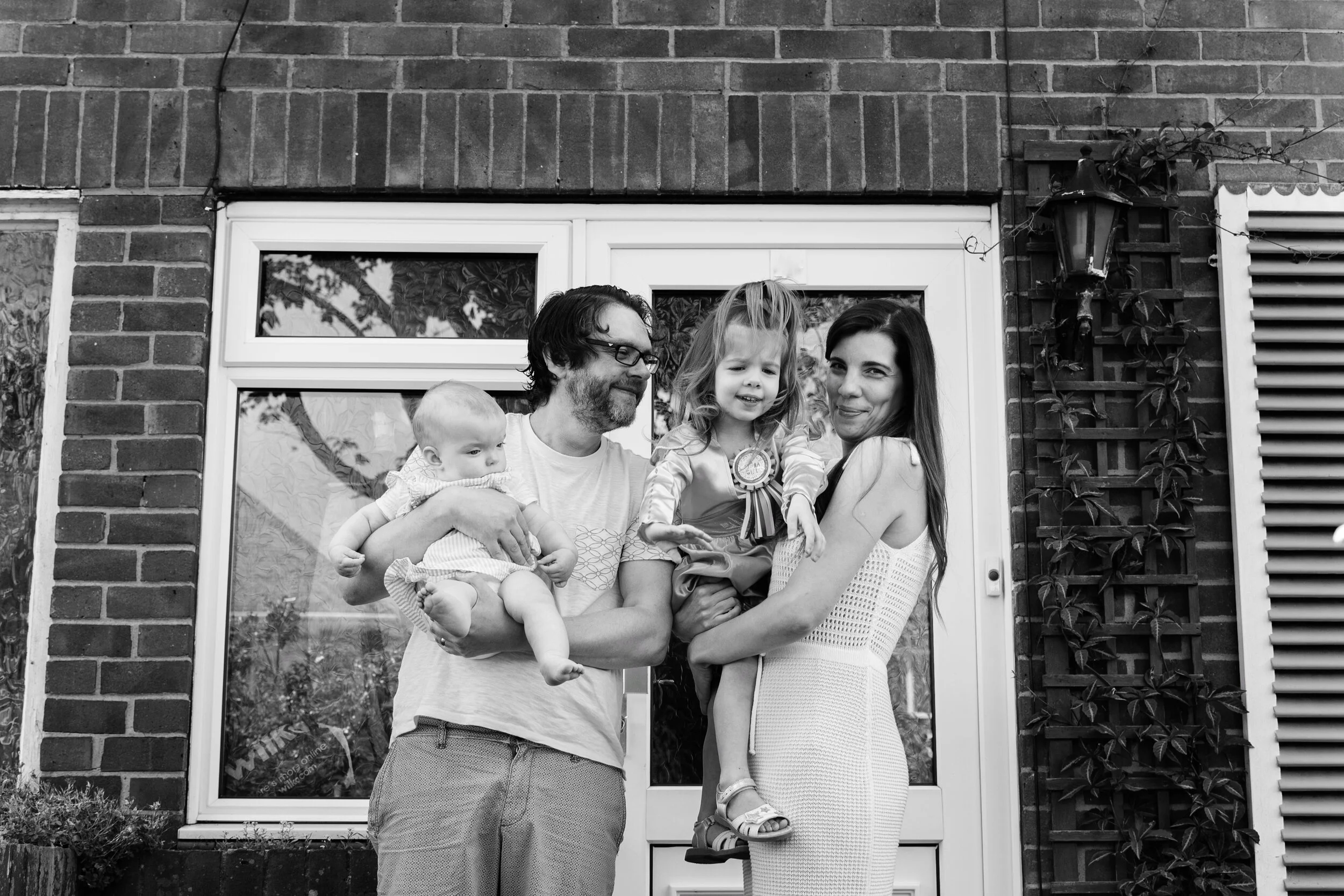 A black and white photo of a family with two adults holding two young children in front of a brick house with windows and plants.