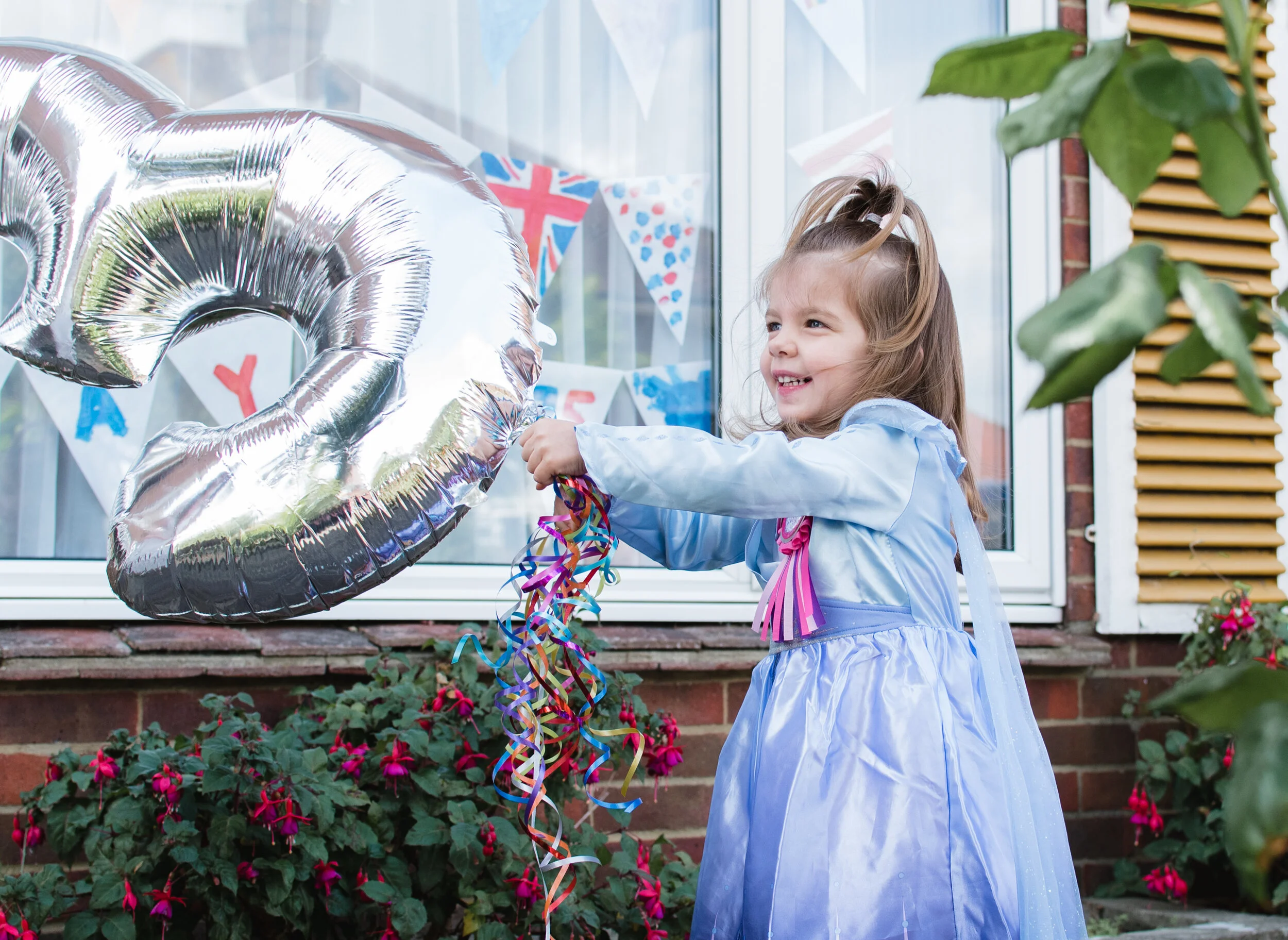 Young girl in purple dress holding large silver number 5 balloon with colorful ribbons, standing outside with festive decorations and plants.