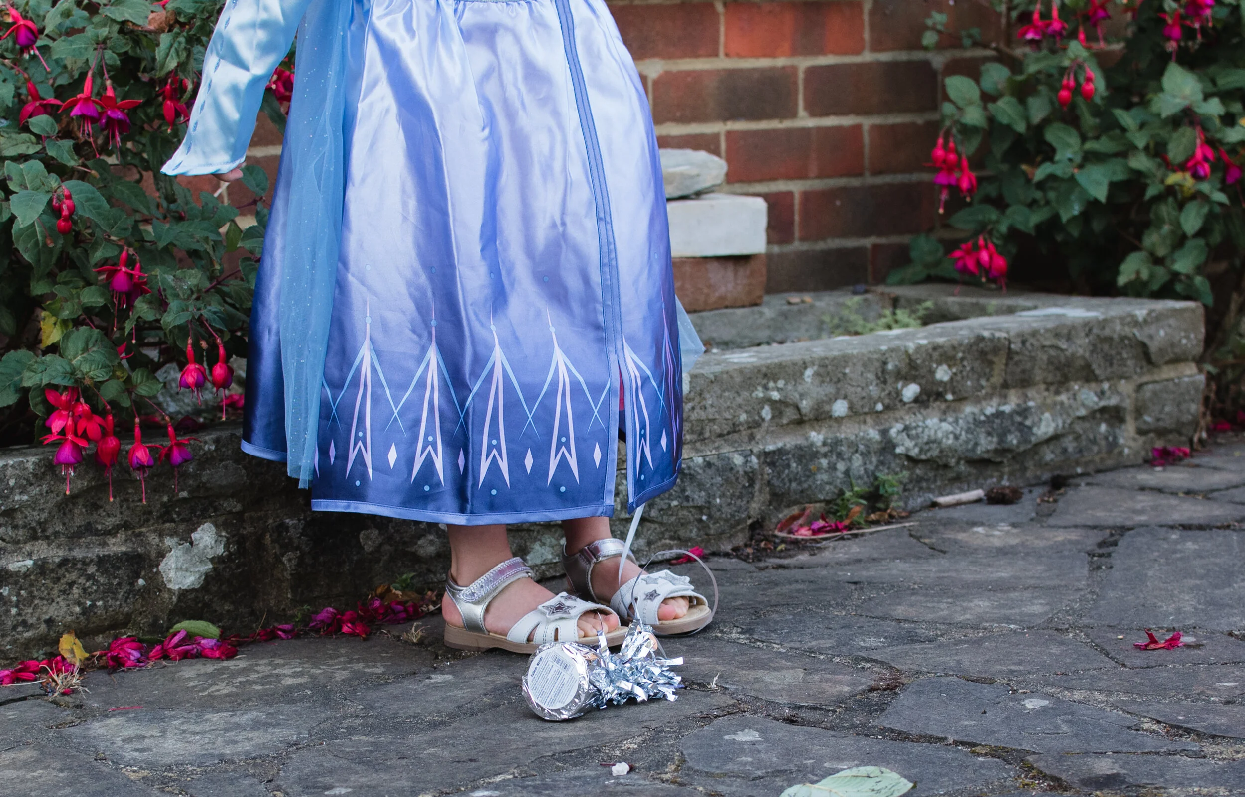 Close-up of a child in a blue dress and silver sandals standing on a stone path, with a balloon weight and pink flowers nearby.