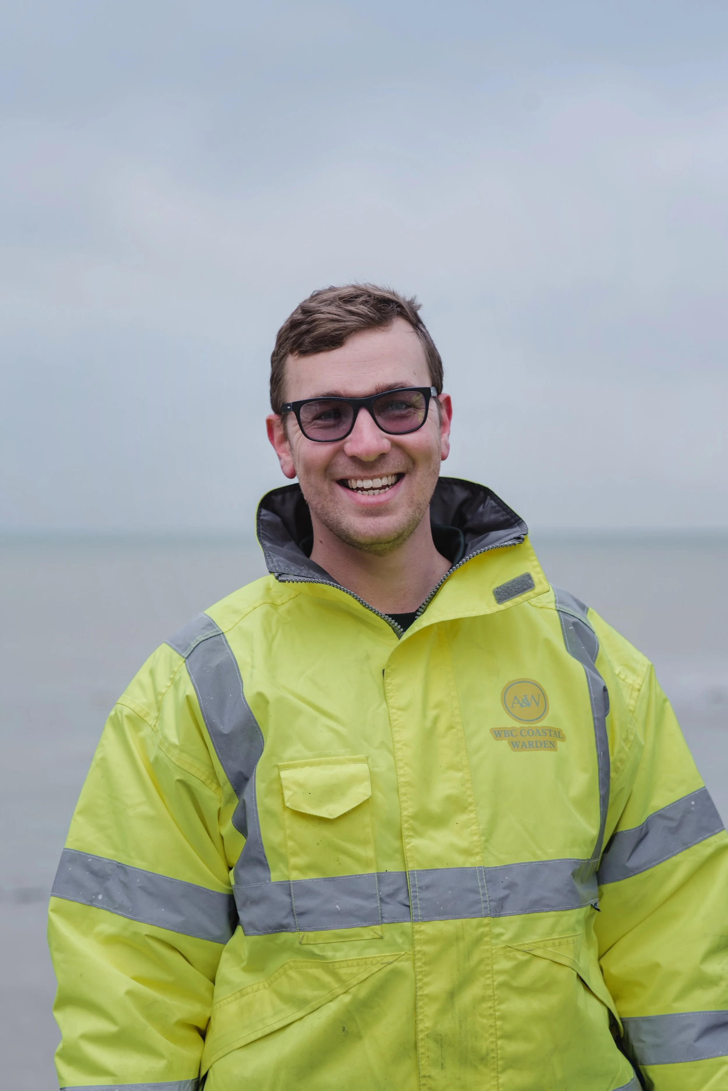 Person wearing a yellow reflective jacket with glasses and smiling, standing outdoors with a cloudy sky.