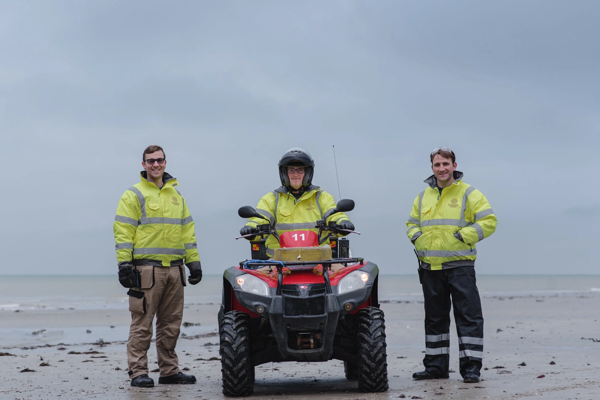 Three people in high-visibility jackets standing on a beach, with one sitting on an ATV, cloudy sky in the background.