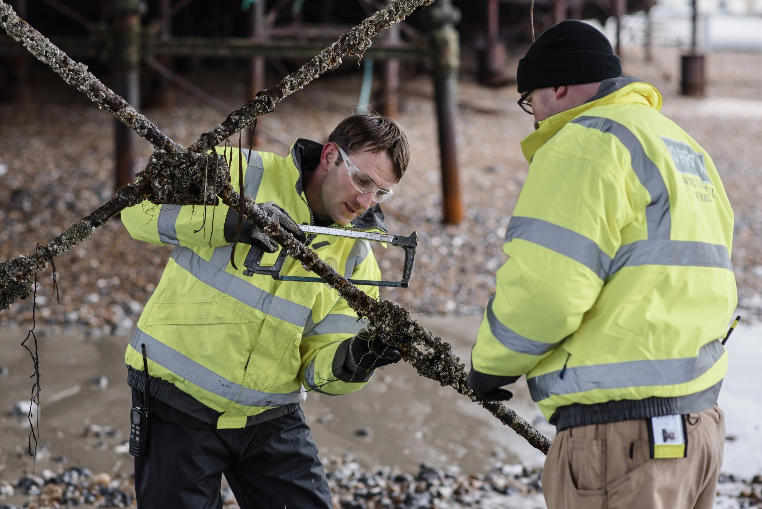 Two workers in yellow safety jackets inspecting a barnacle-covered metal structure on a beach.