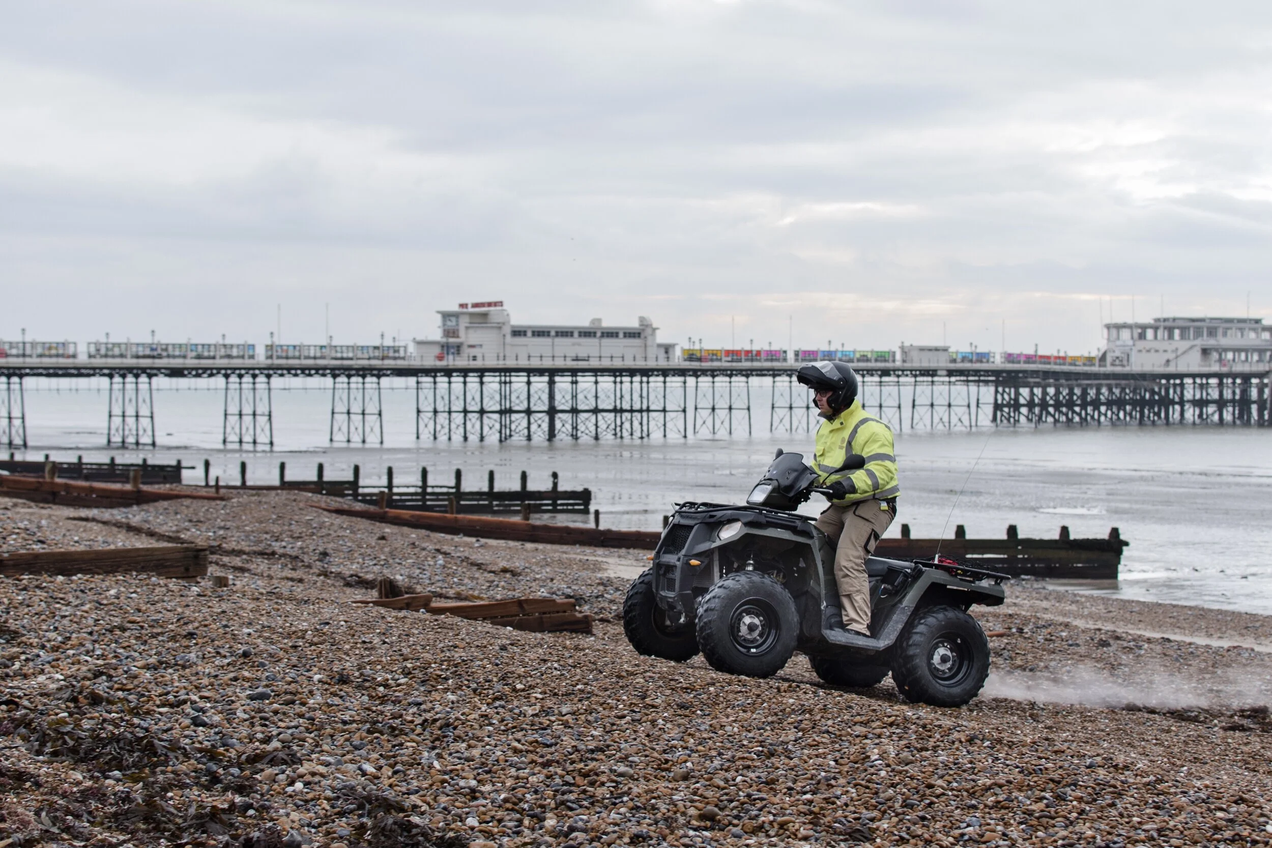 Person on ATV riding on a pebble beach with a pier and overcast sky in the background.