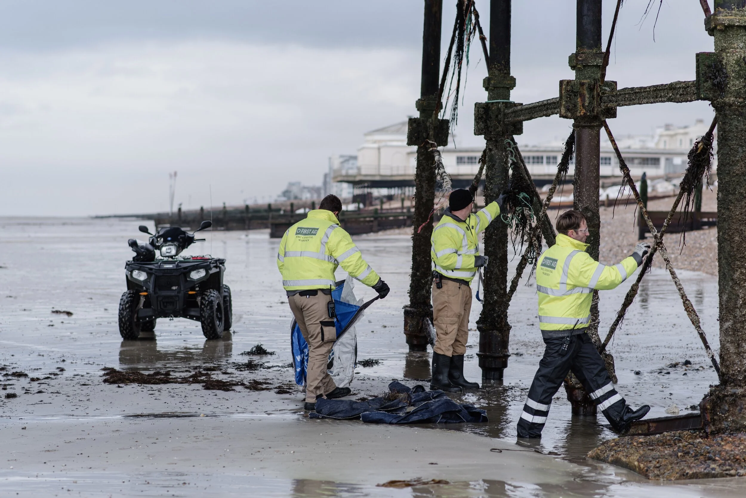 Three workers in high-visibility jackets inspecting and cleaning a seaside pier structure, with an ATV parked nearby on a wet, sandy beach.