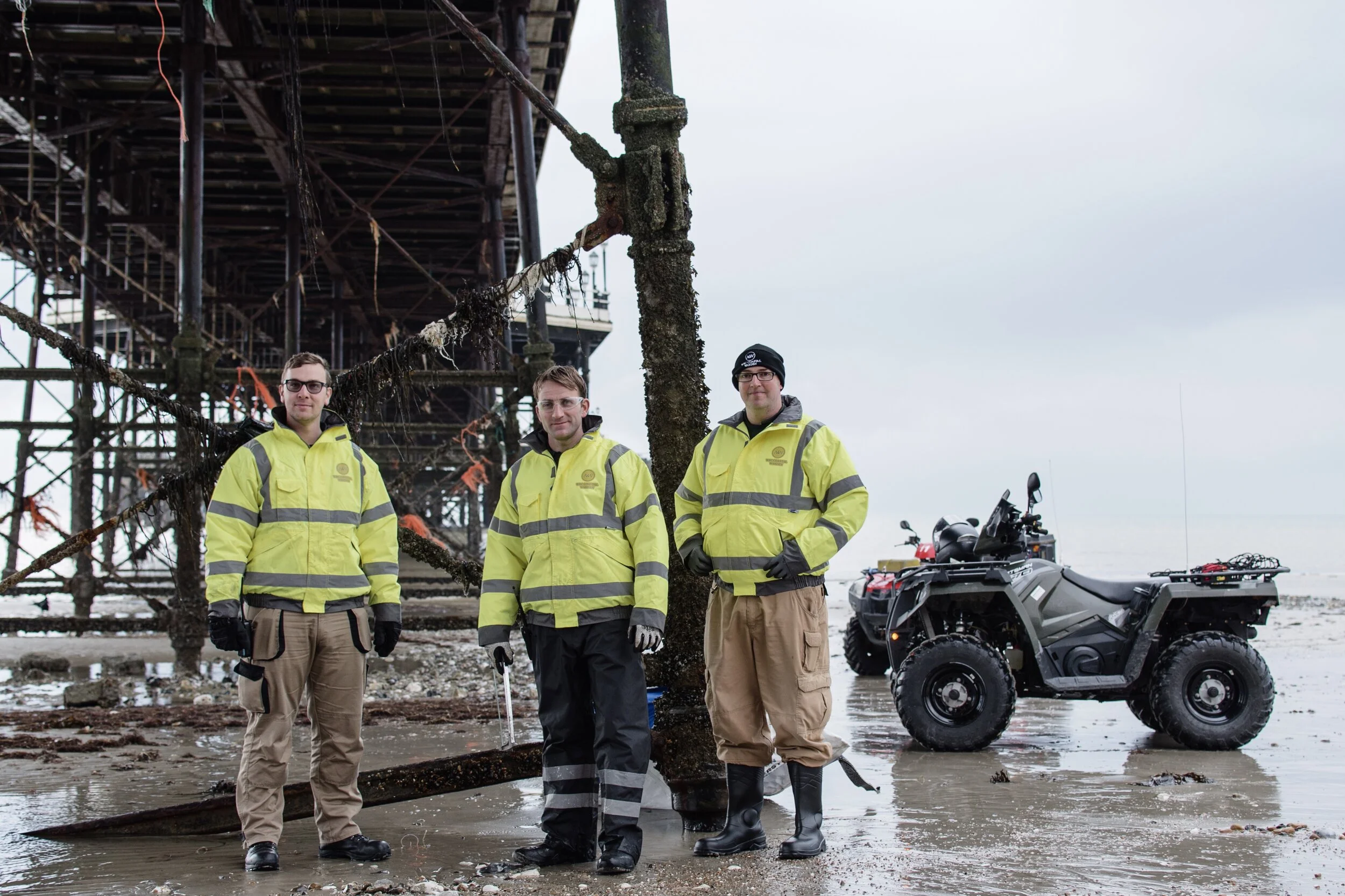 Three workers in yellow reflective jackets standing on a beach under a pier next to an ATV.