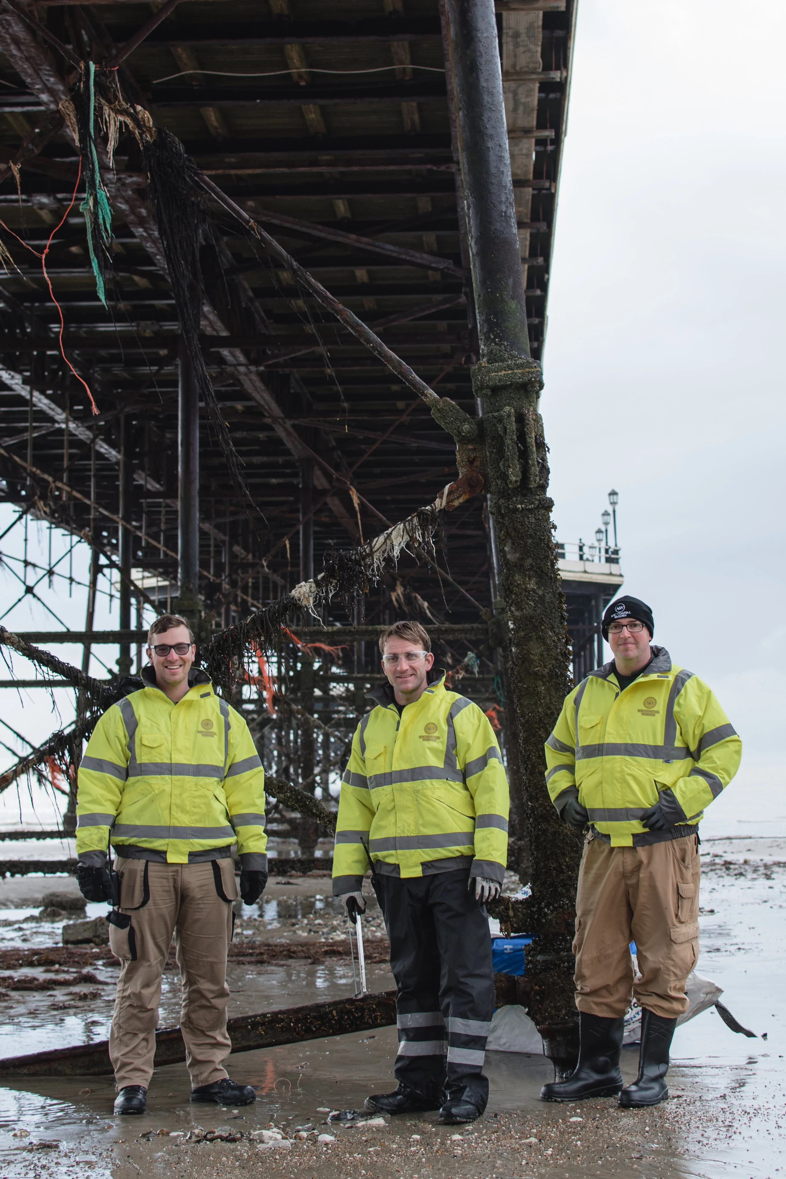 Three people in high-visibility jackets standing under a pier, with exposed beams and seaweed on the beach.