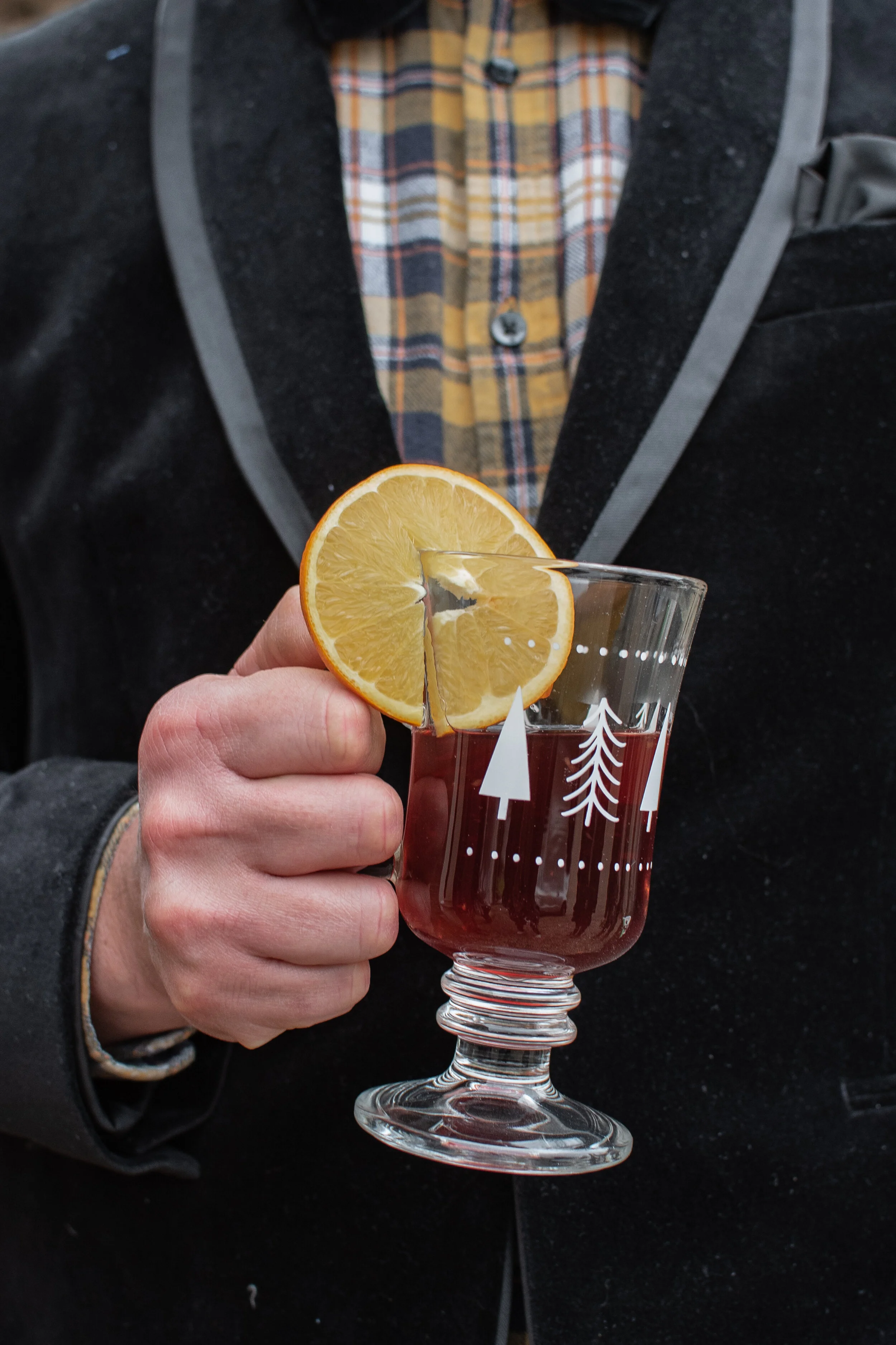 Person holding a festive glass mug filled with a dark beverage and a lemon slice, wearing a velvet jacket and plaid shirt.