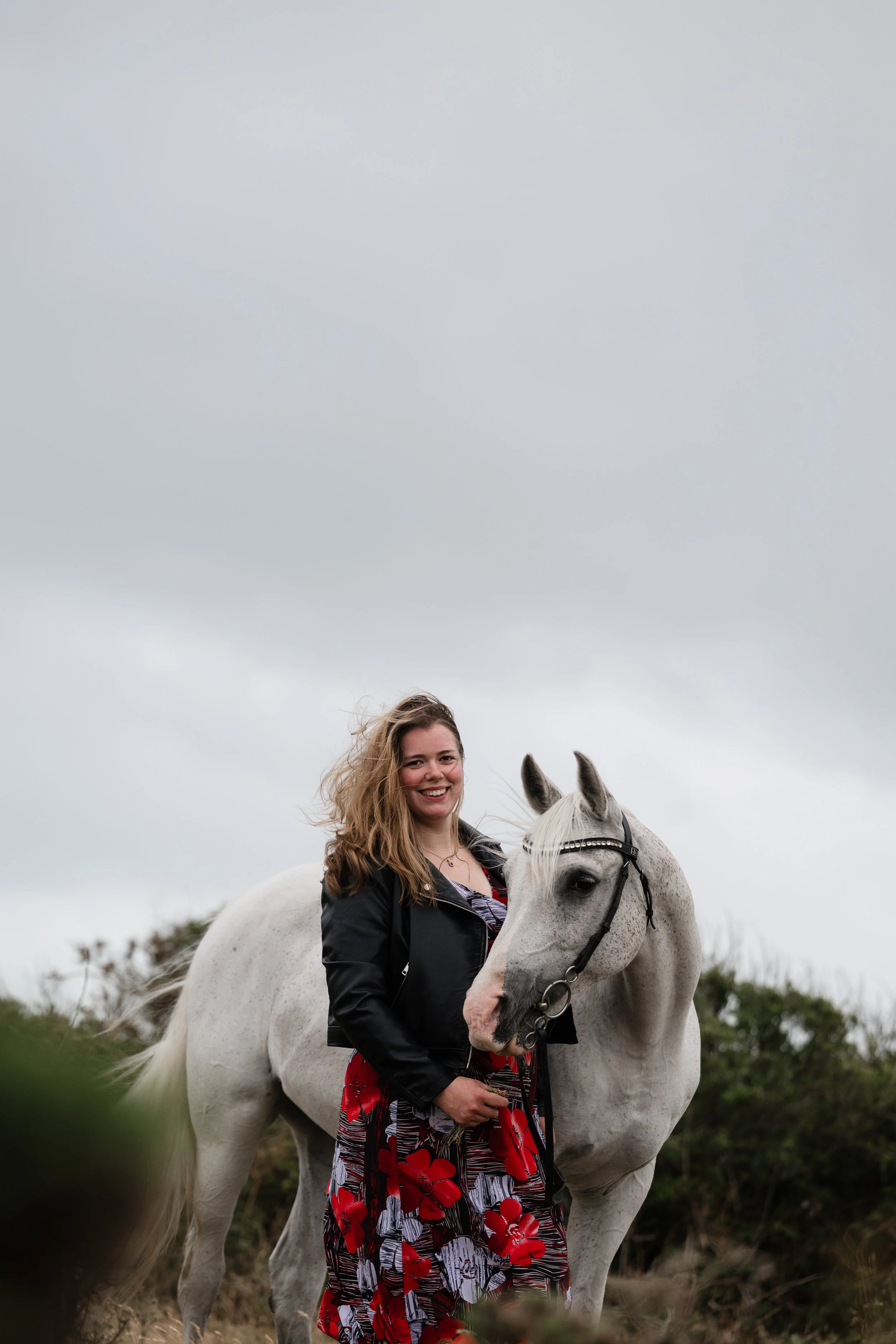 A woman with curly hair, wearing a red floral dress and black jacket, is smiling and sitting on a white horse outdoors on a cloudy day.