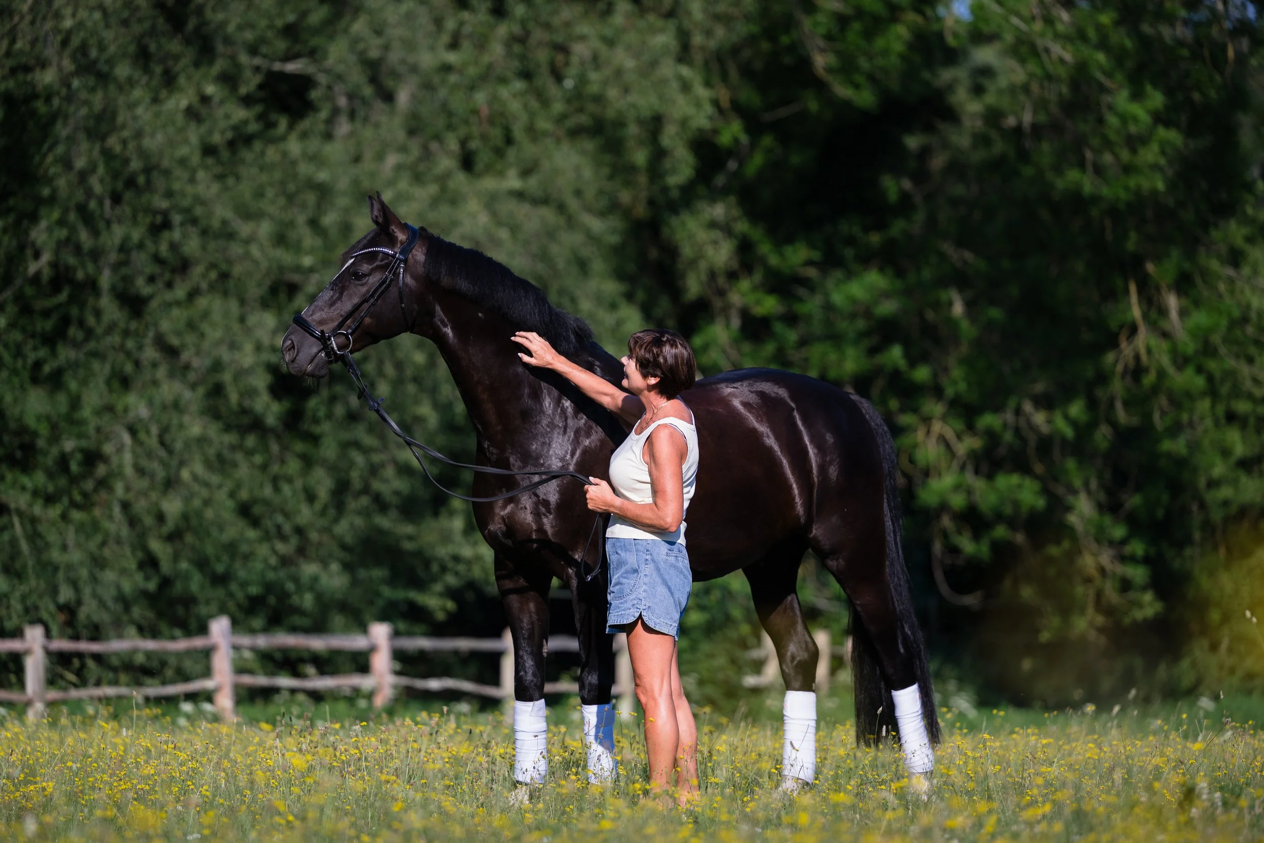 A woman in casual clothing standing next to a dark brown horse with white socks, petting its neck outdoors with green trees in the background.