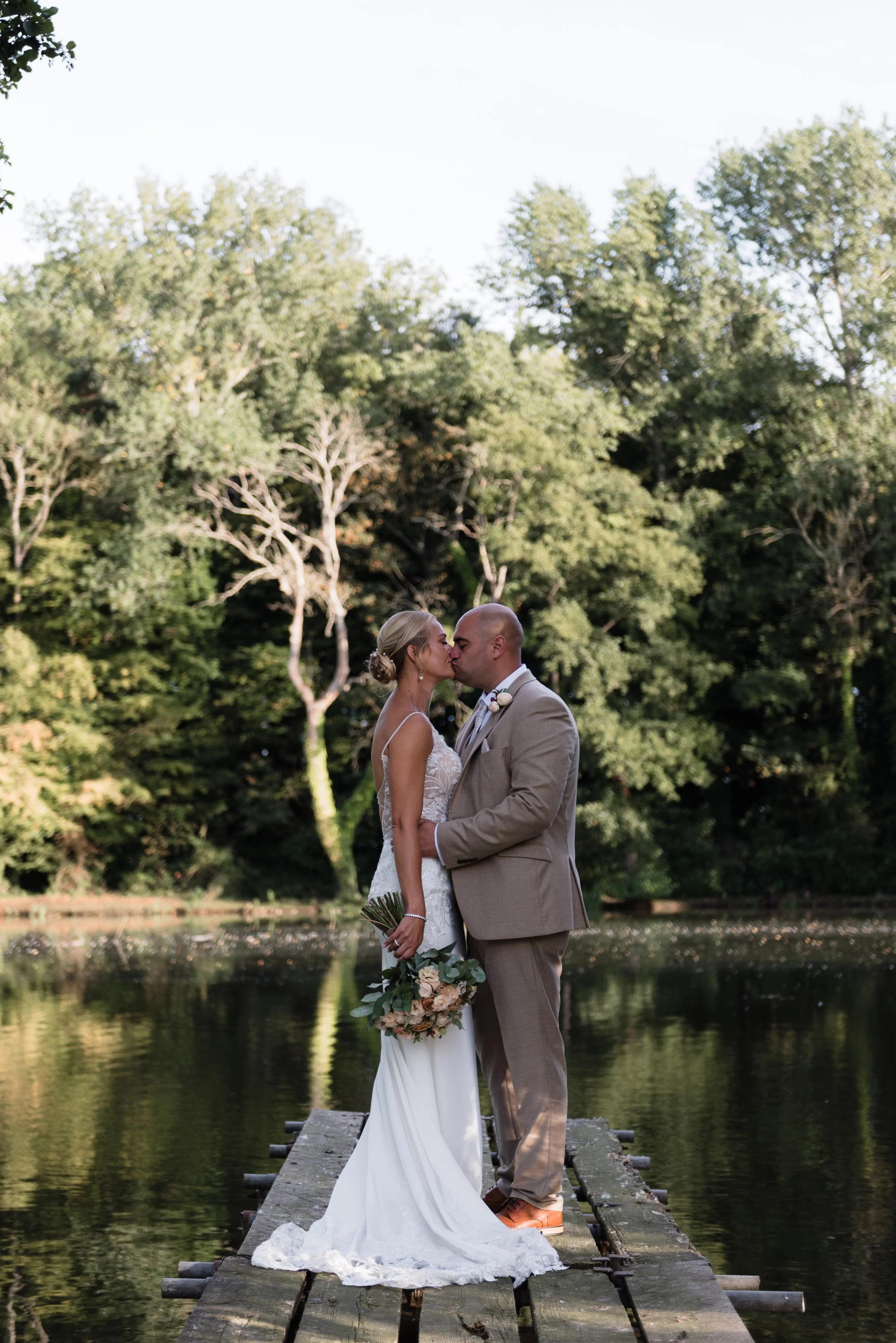 A newlywed couple sharing a kiss in front of a lake with the bride holding a bouquet, during daytime.