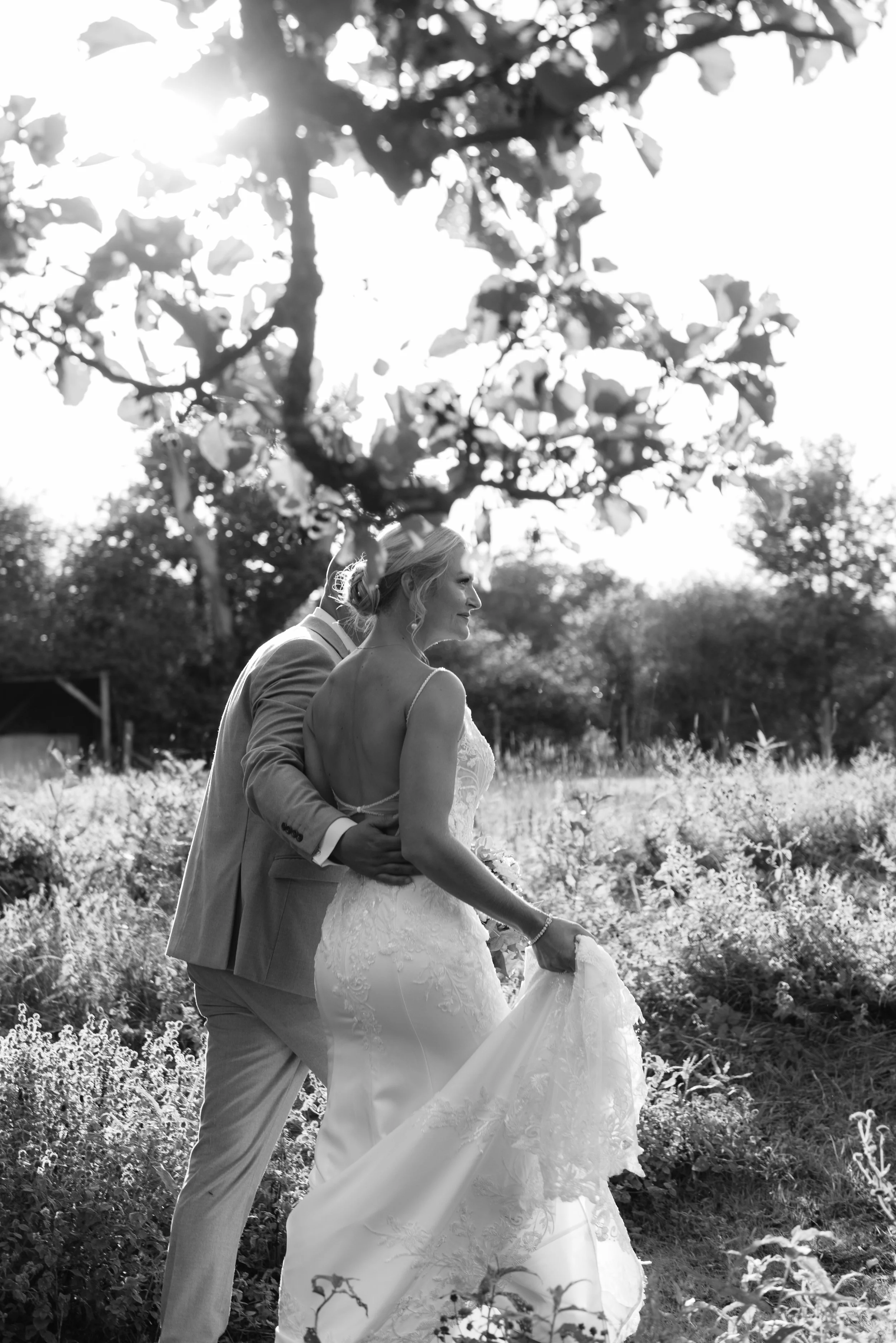 Black and white photo of a bride and groom walking through long grass backlit by low sun