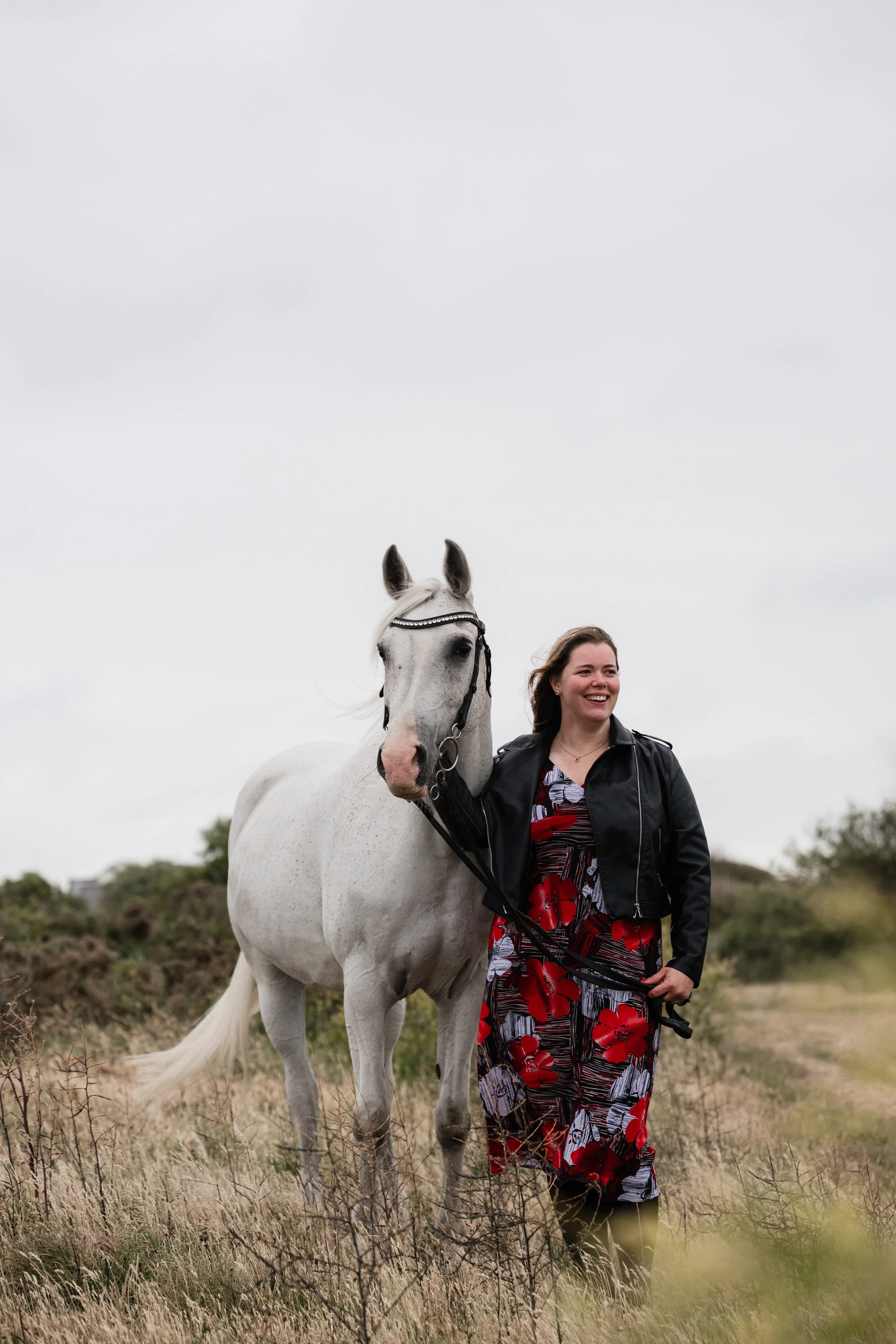 A woman standing outdoors on a grassy field, smiling, holding a white horse by the reins. The woman is wearing a black leather jacket and a colorful dress with red and black patterns. The sky is overcast, and the background is blurred with some trees
