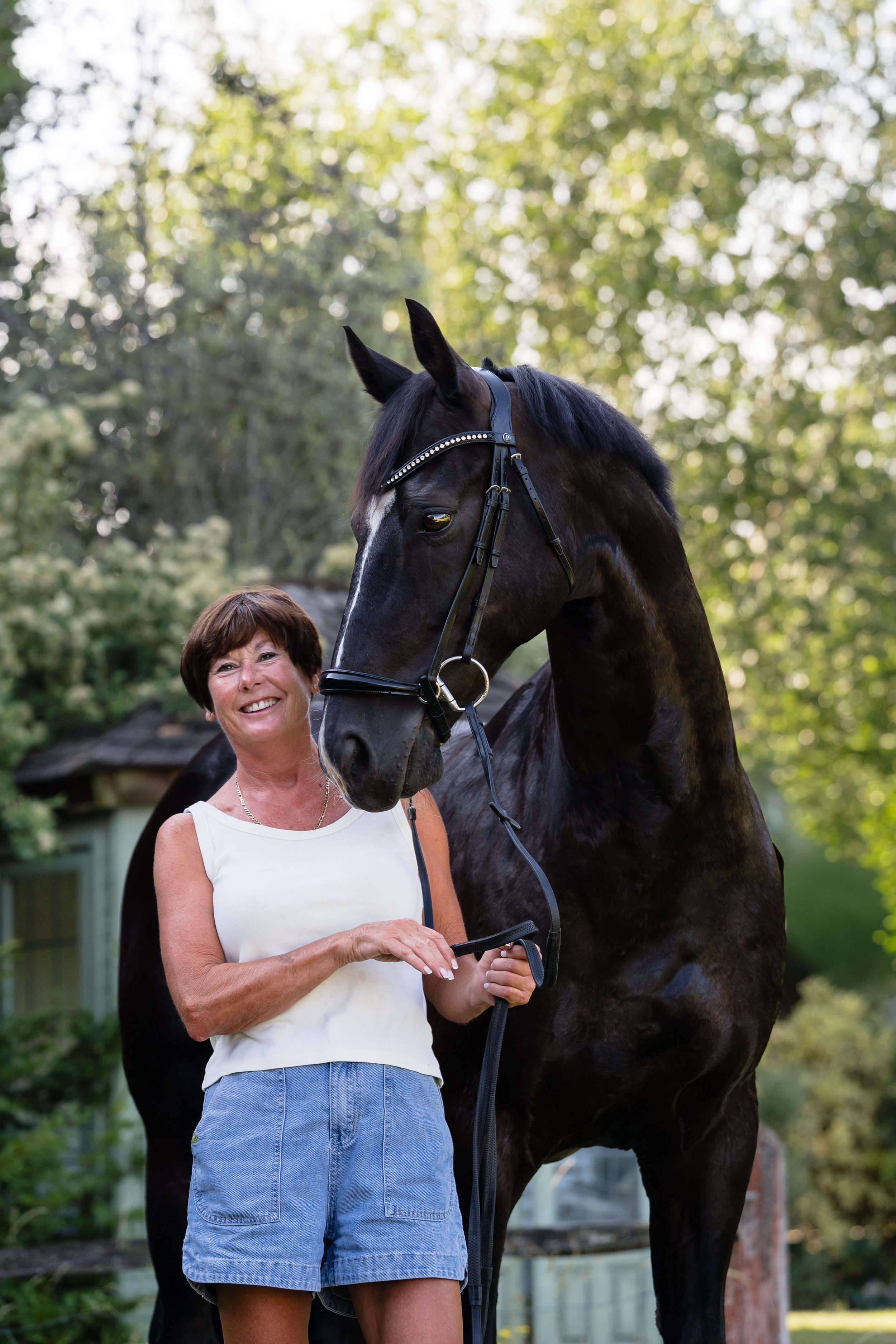 A woman standing outdoors holding a black horse with a decorative bridle. The woman is smiling and wearing a white sleeveless top and denim shorts. The background features trees and a house.