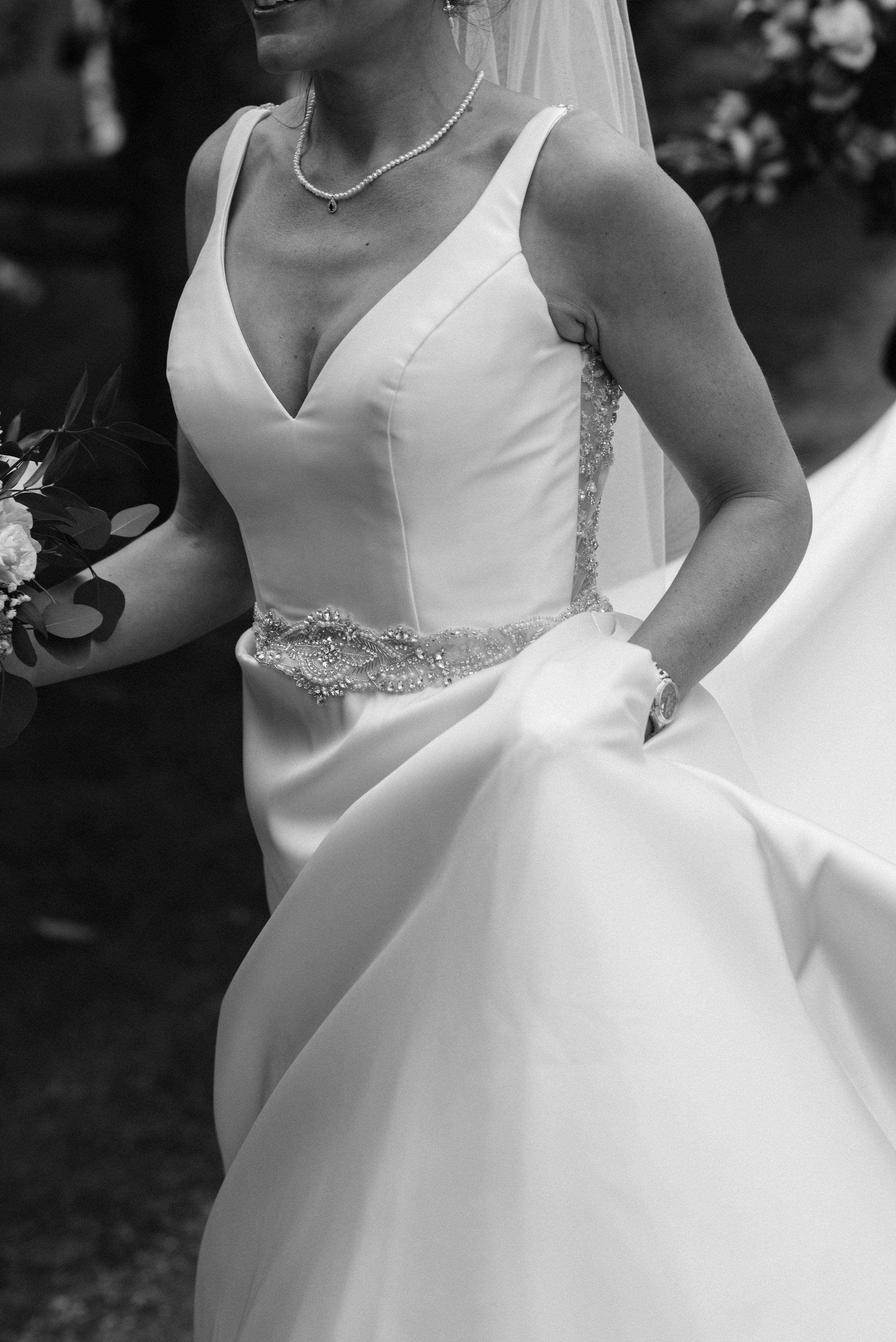 Black and white photo of a bride in a wedding dress, holding flowers, with intricate beading and lace details on the gown.