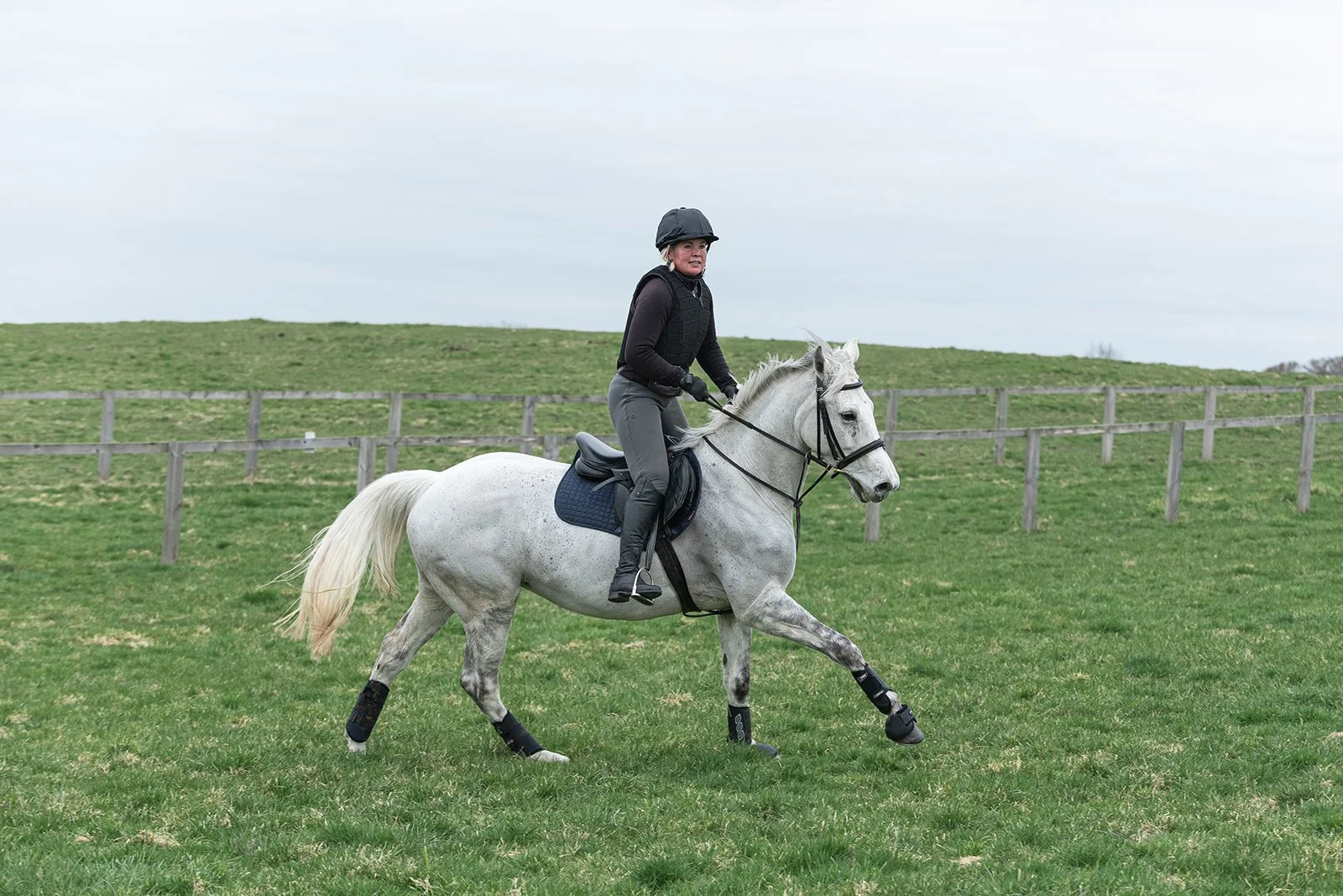 Person riding a grey horse in a grassy field with a wooden fence.