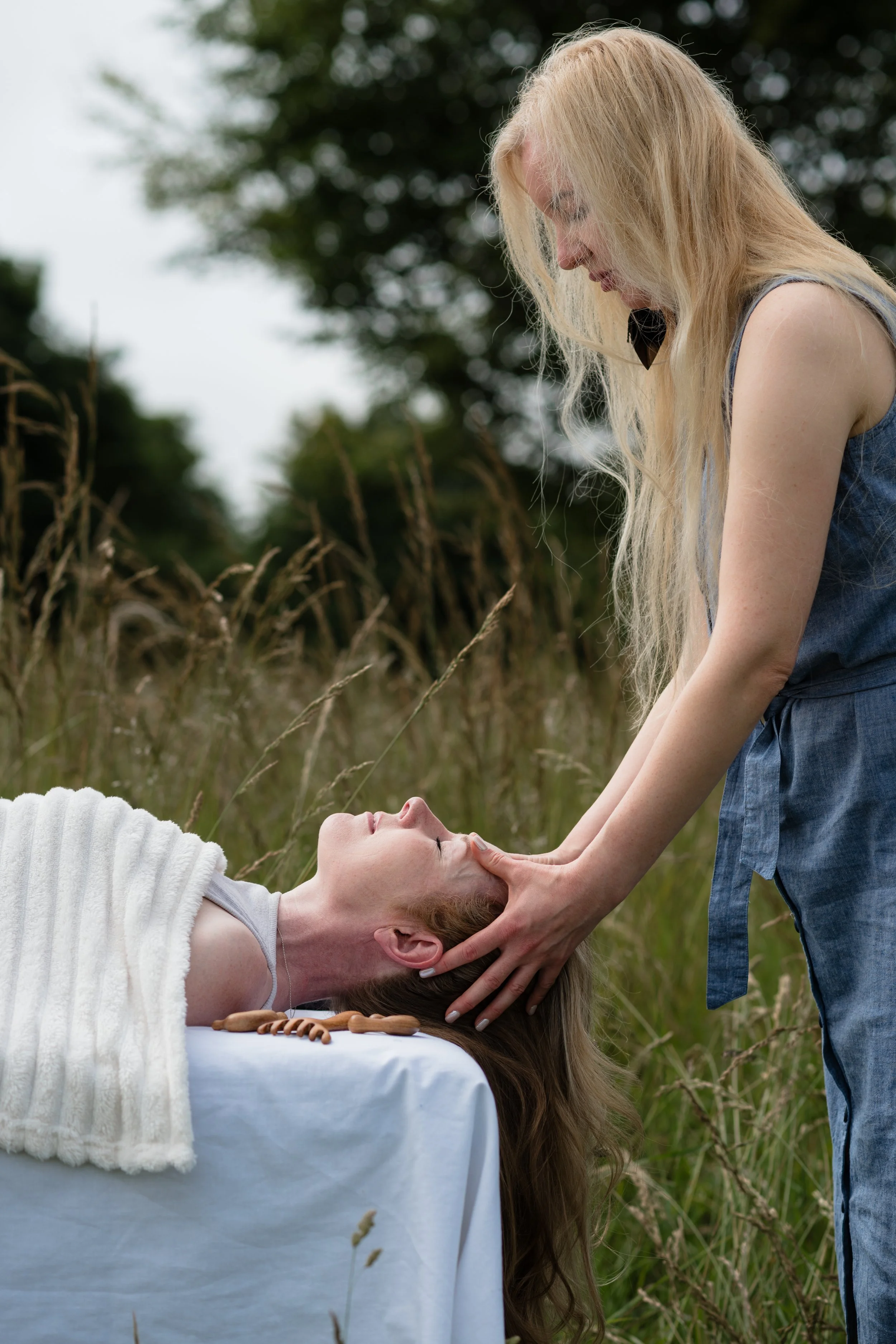 A woman laying on a table with a white cloth, receiving a healing blessing from another woman with long blonde hair, in an outdoor natural setting with tall grass and trees in the background.
