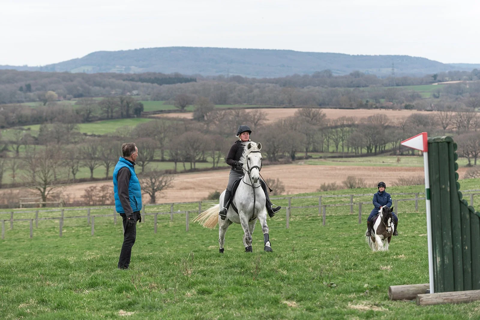 Two people horseback riding in a rural field while another person stands nearby observing. Hills and trees in the background.