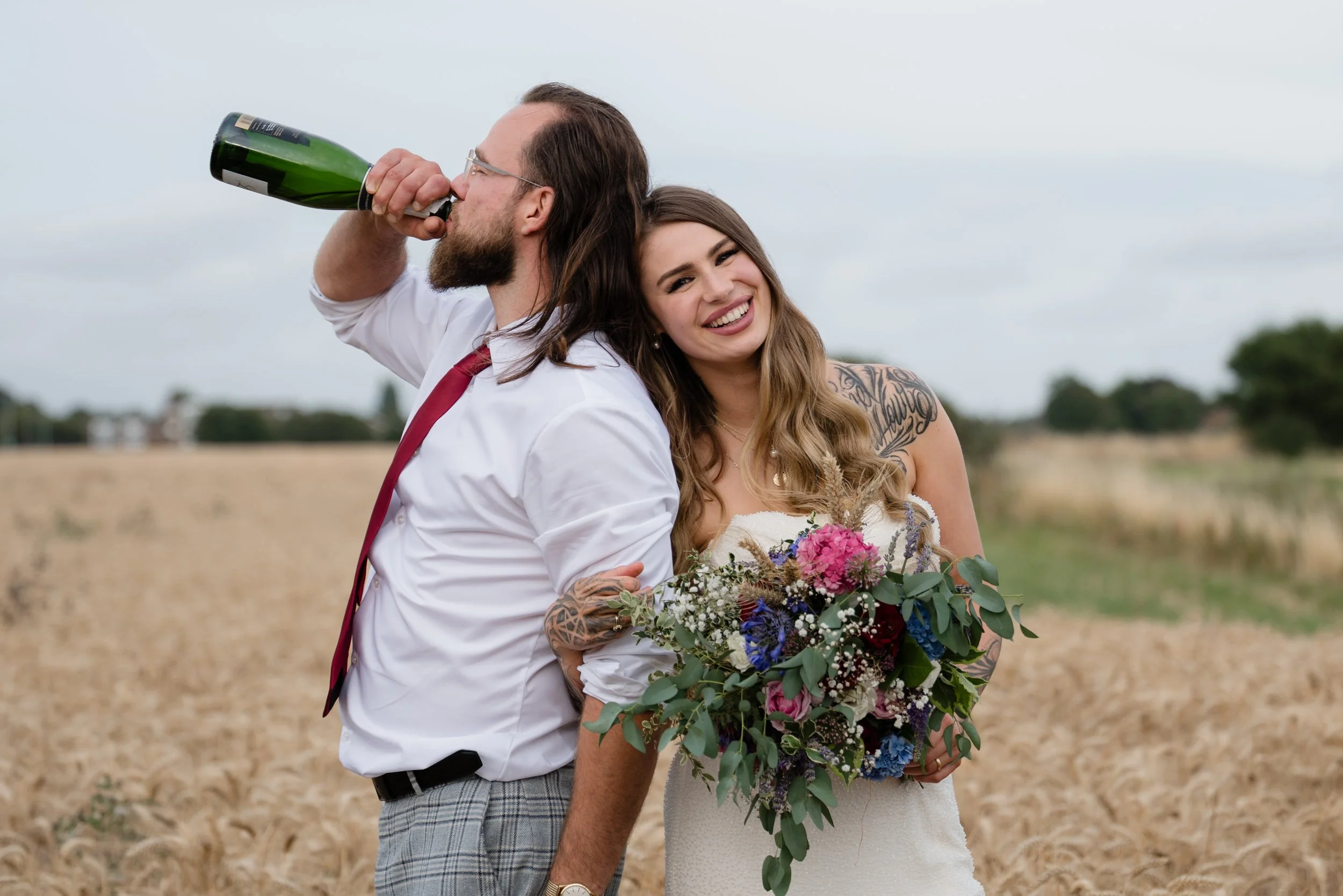 bride and groom in a rural setting holding a bouquet and drinking champagne