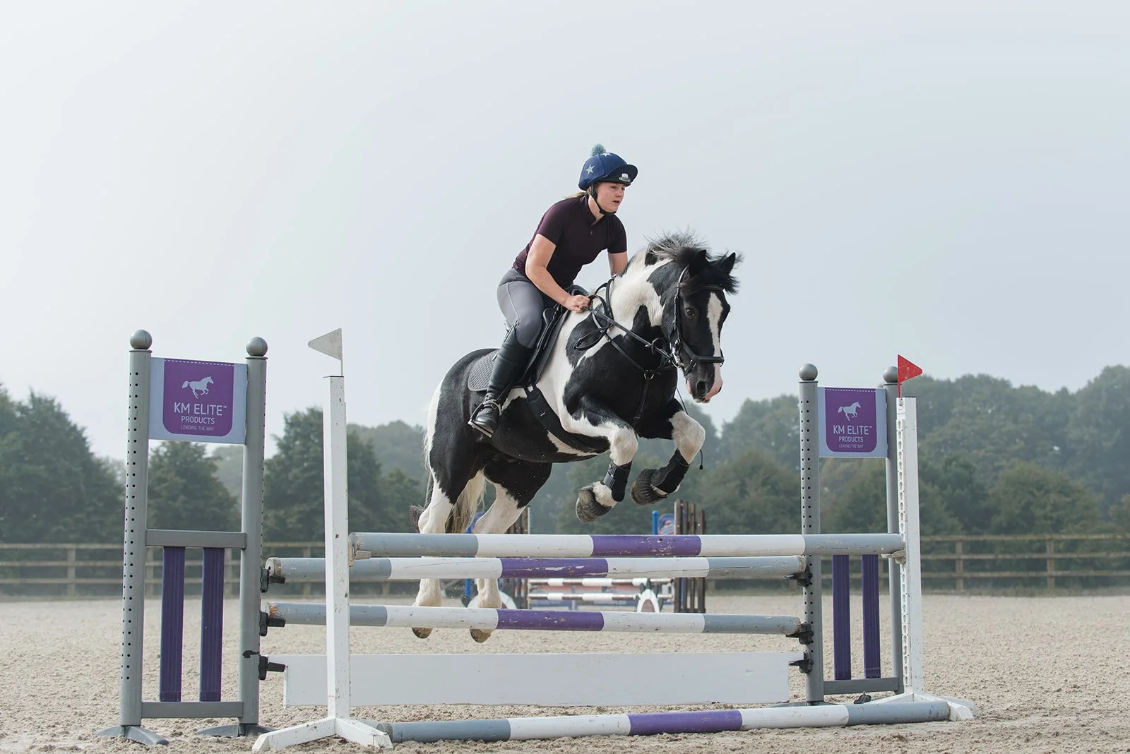 A person riding a piebald horse, jumping over a hurdle in an equestrian event. The setting is an outdoor arena with trees in the background. "KM Elite Products" is visible on the jump poles.