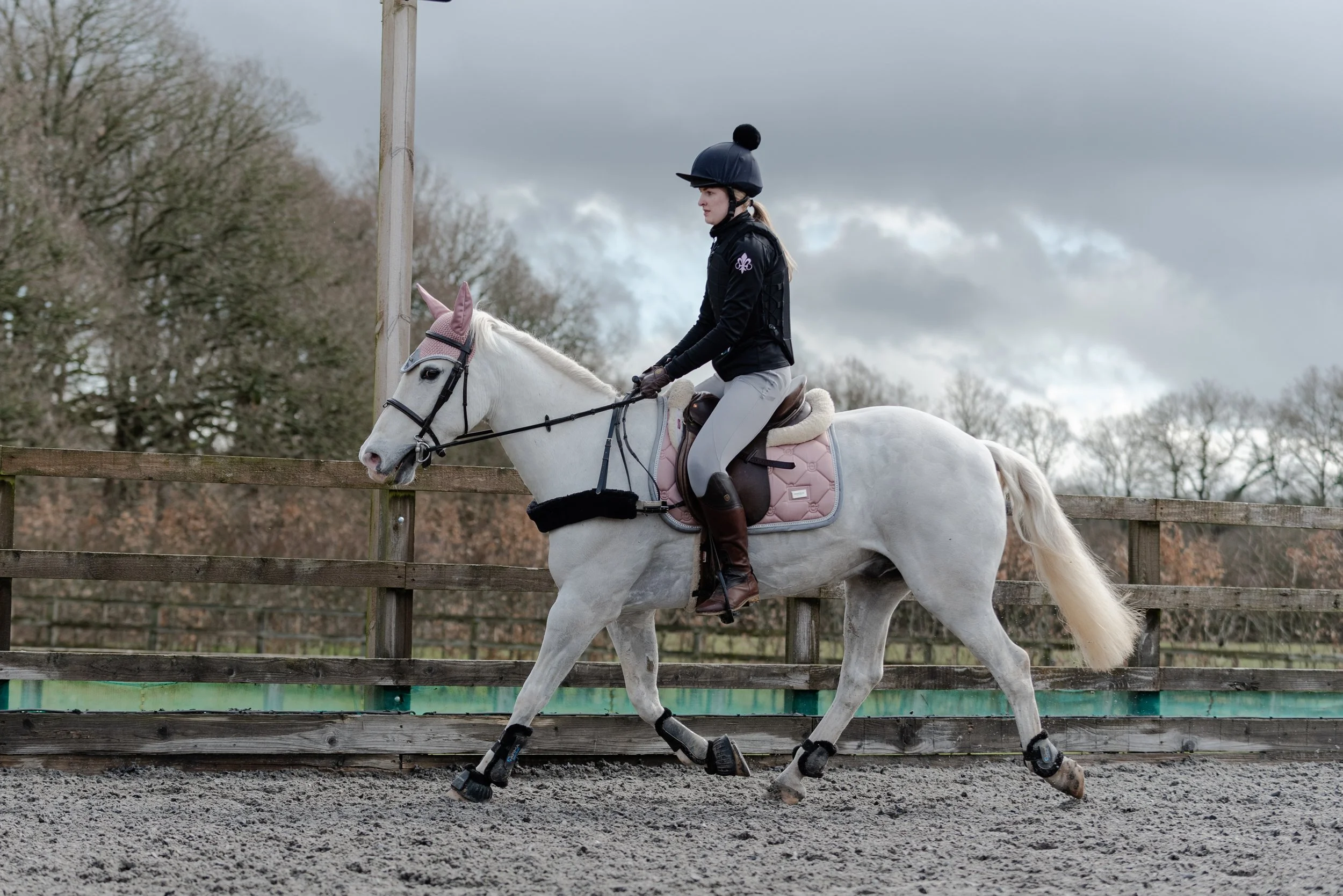 A person riding a grey horse in an outdoor equestrian arena, wearing a helmet and equestrian attire, with a cloudy sky in the background.
