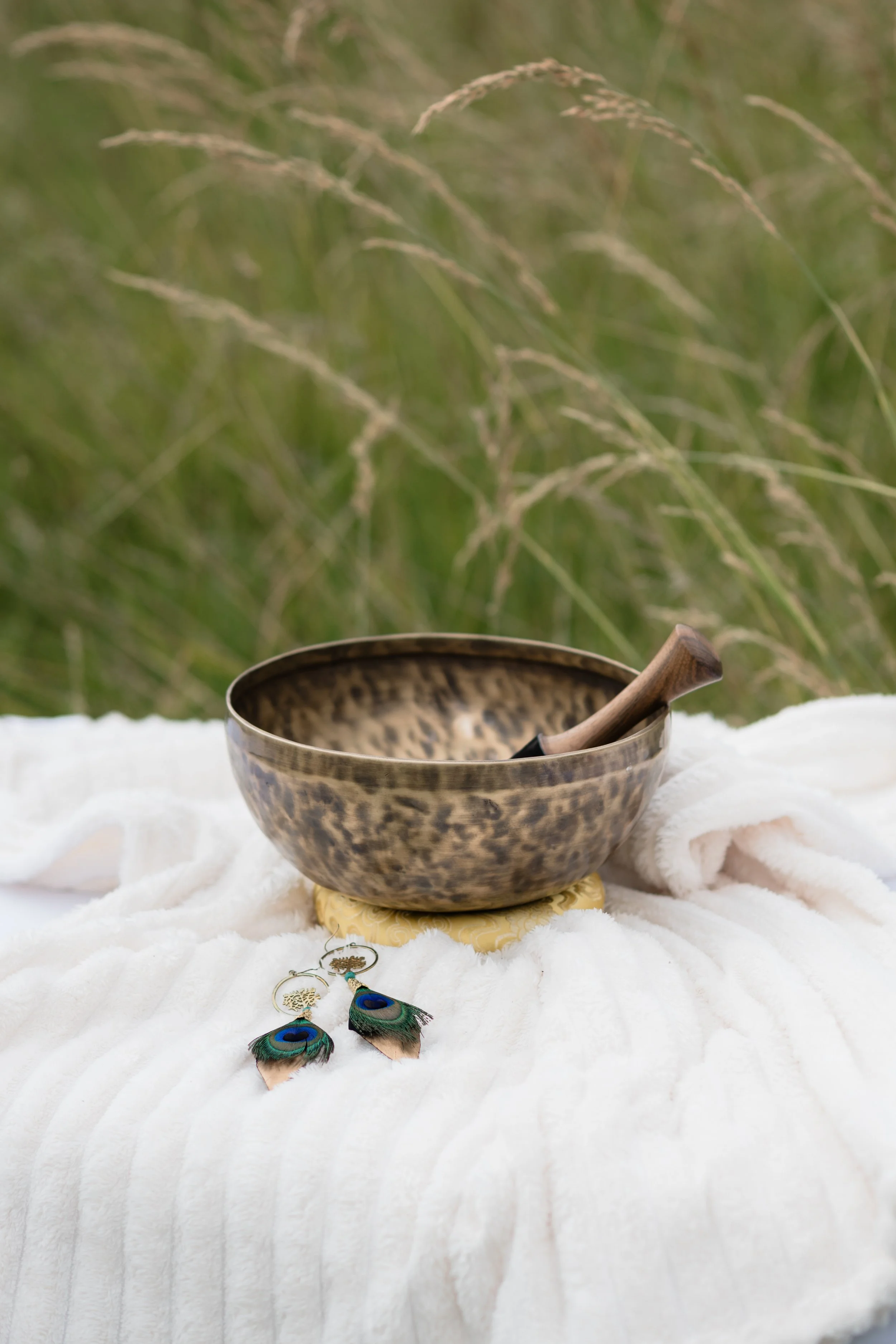 A metallic singing bowl with a wooden striker resting on it, placed on a white textured cloth with peacock feather earrings nearby. In the background, there is green grass and plants.