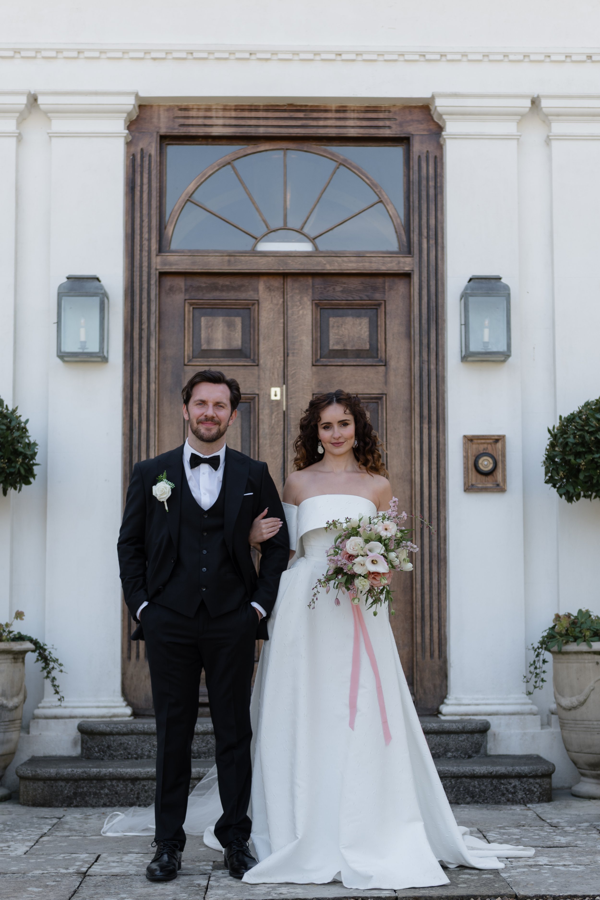 Bride and groom standing in front of a wooden door, bride holding a bouquet, groom in a black tuxedo, wedding setting.