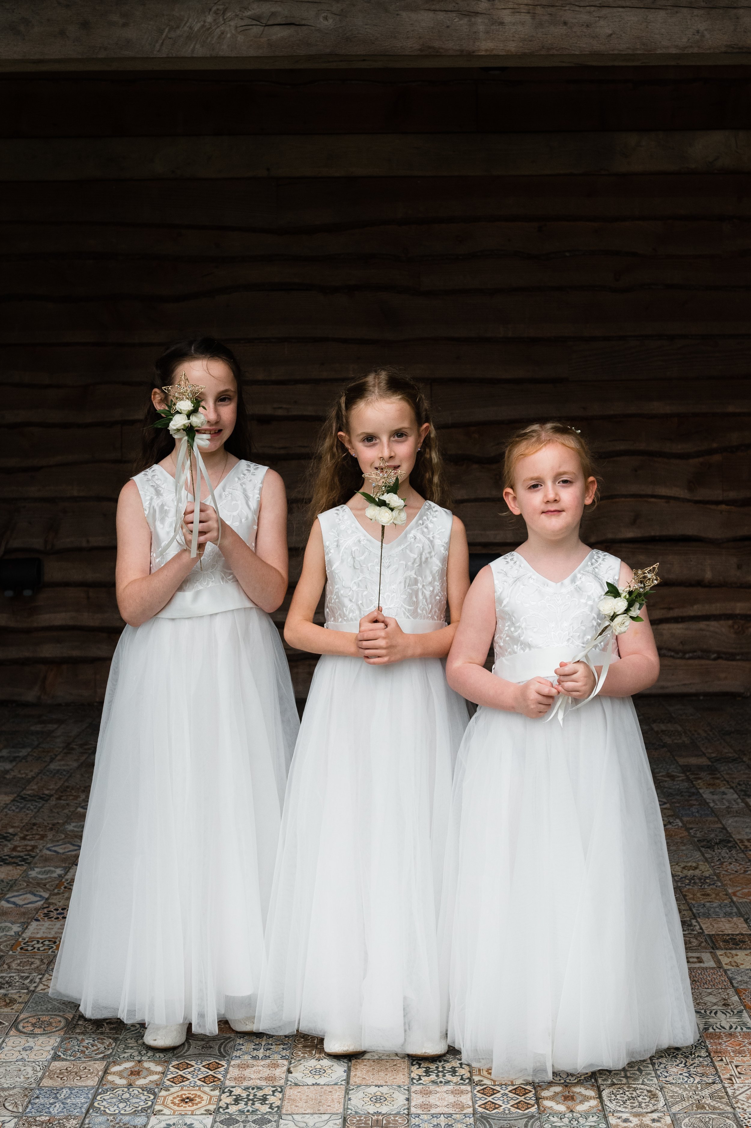 Three young girls in white dresses holding small bouquets, standing indoors on a patterned tile floor with a wooden background.