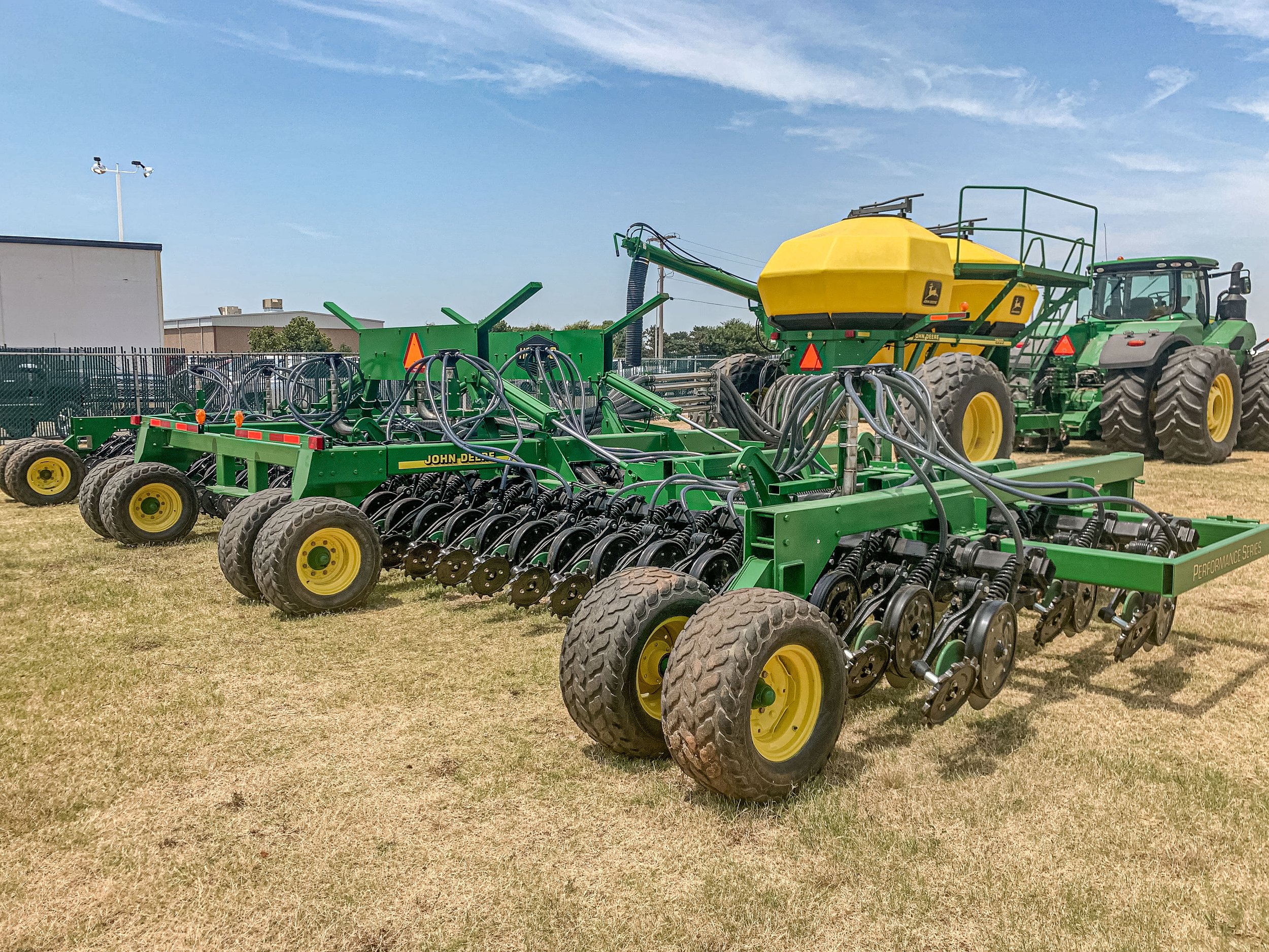 Agricultural equipment including a green John Deere planter, yellow tank, and a green tractor on a grassy field under a blue sky.