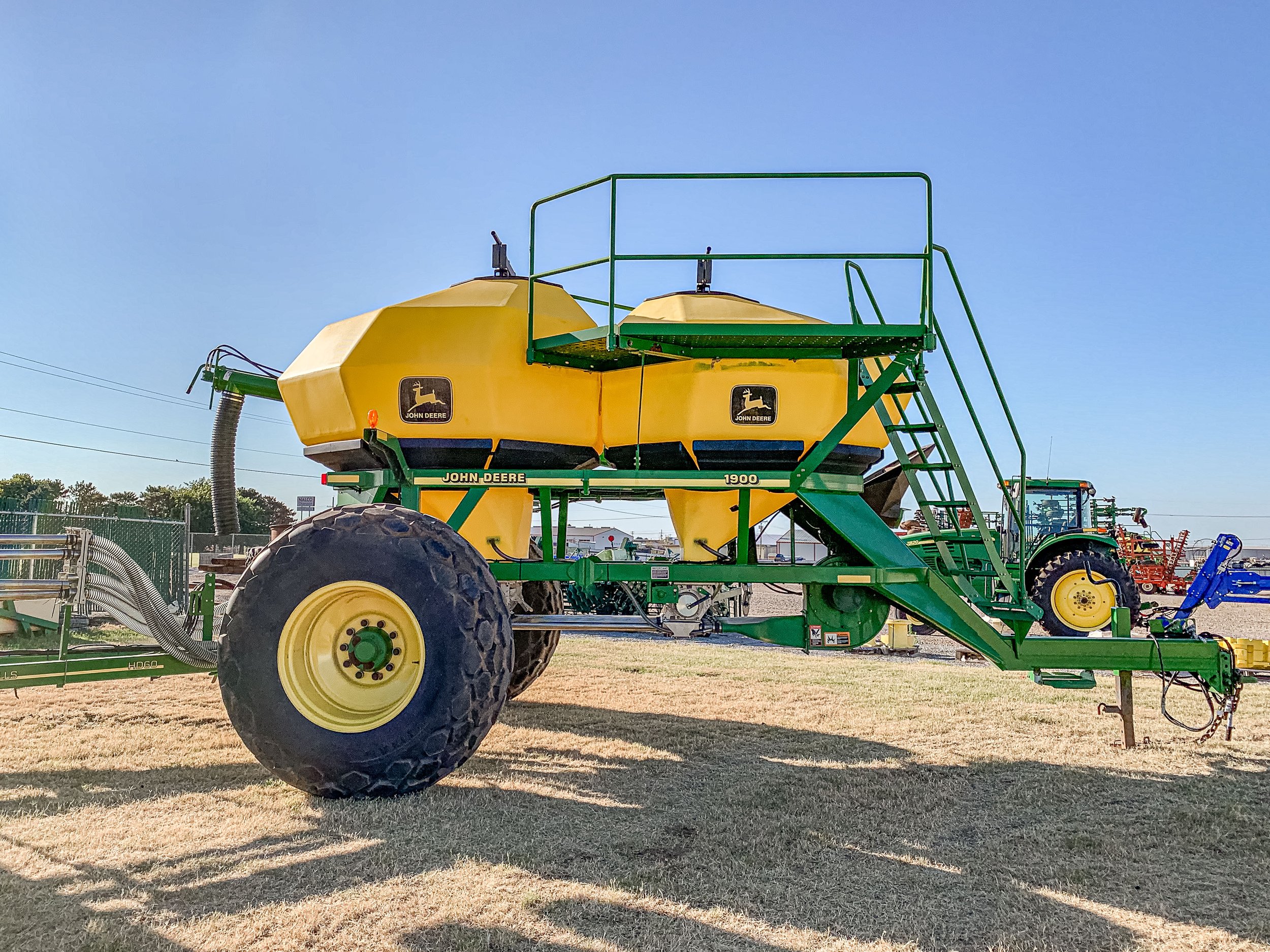A large green and yellow John Deere agricultural sprayer with big tires, set on dry grass with other farming equipment in the background under a clear blue sky.