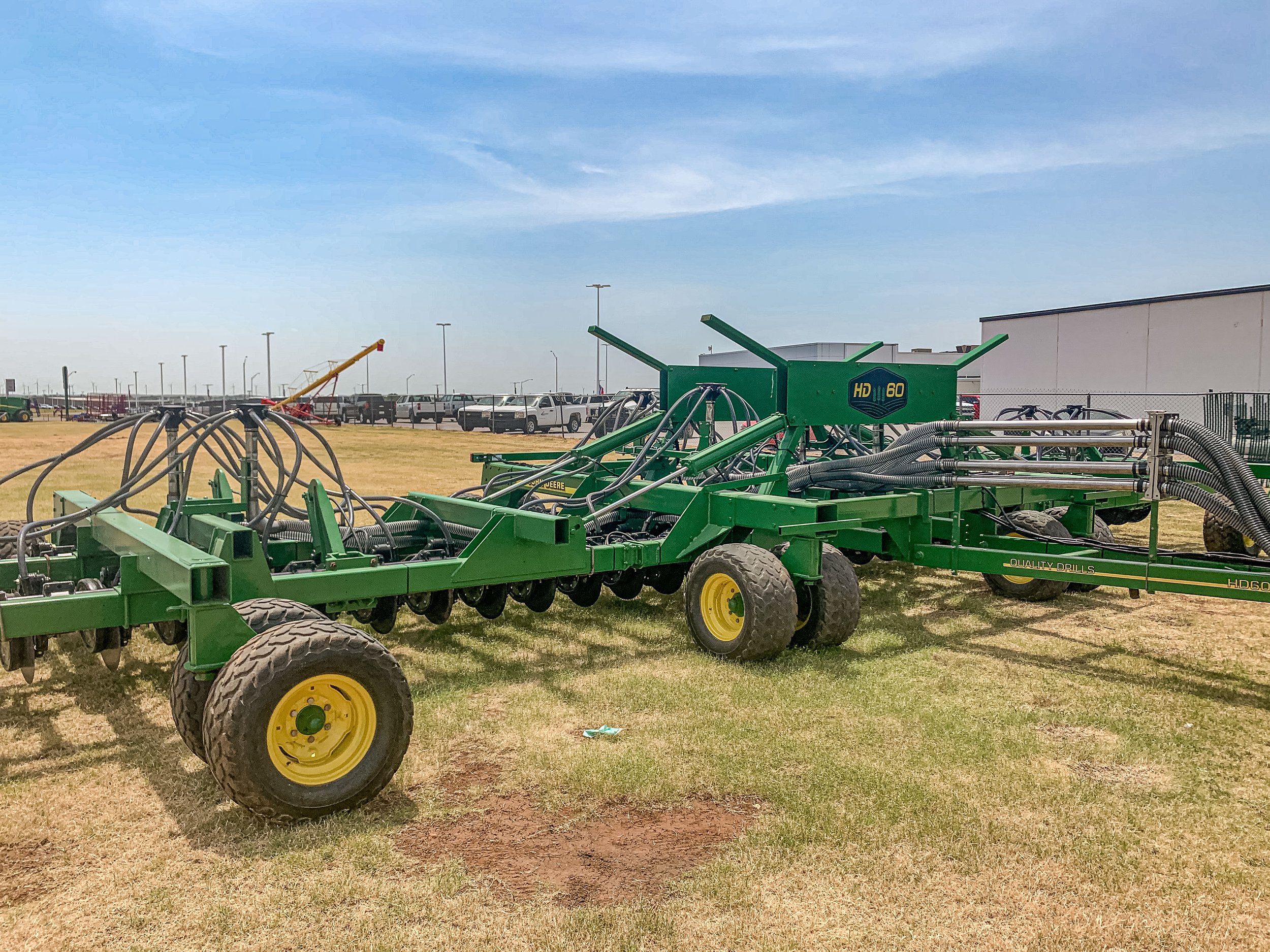 Green John Deere agricultural equipment with large tires and black hoses outdoors on grassy field, with a parking lot and white building in background under blue sky.