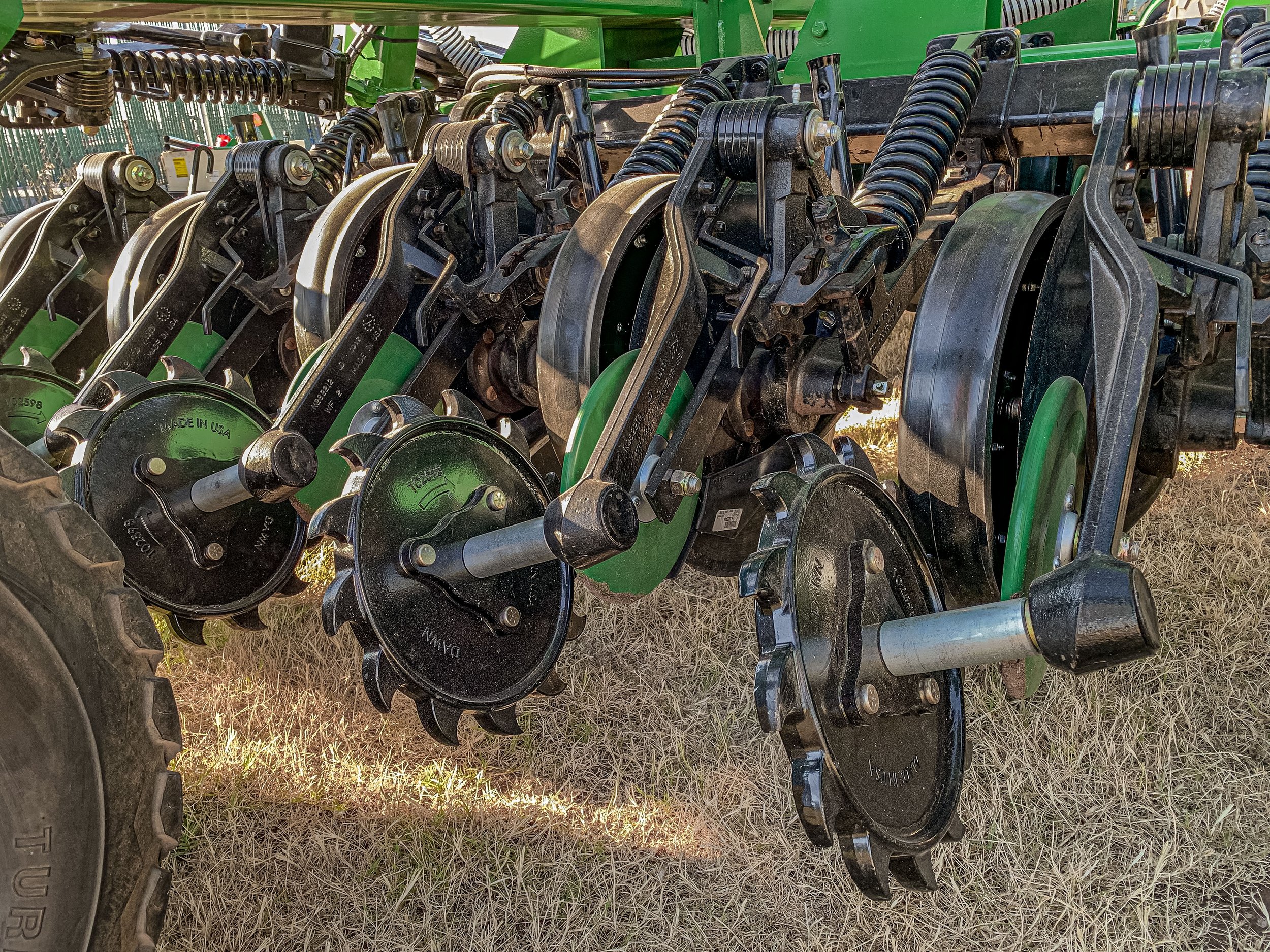 Close-up view of a row of agricultural disc harrow blades attached to a green tractor implement, resting on dry grass.