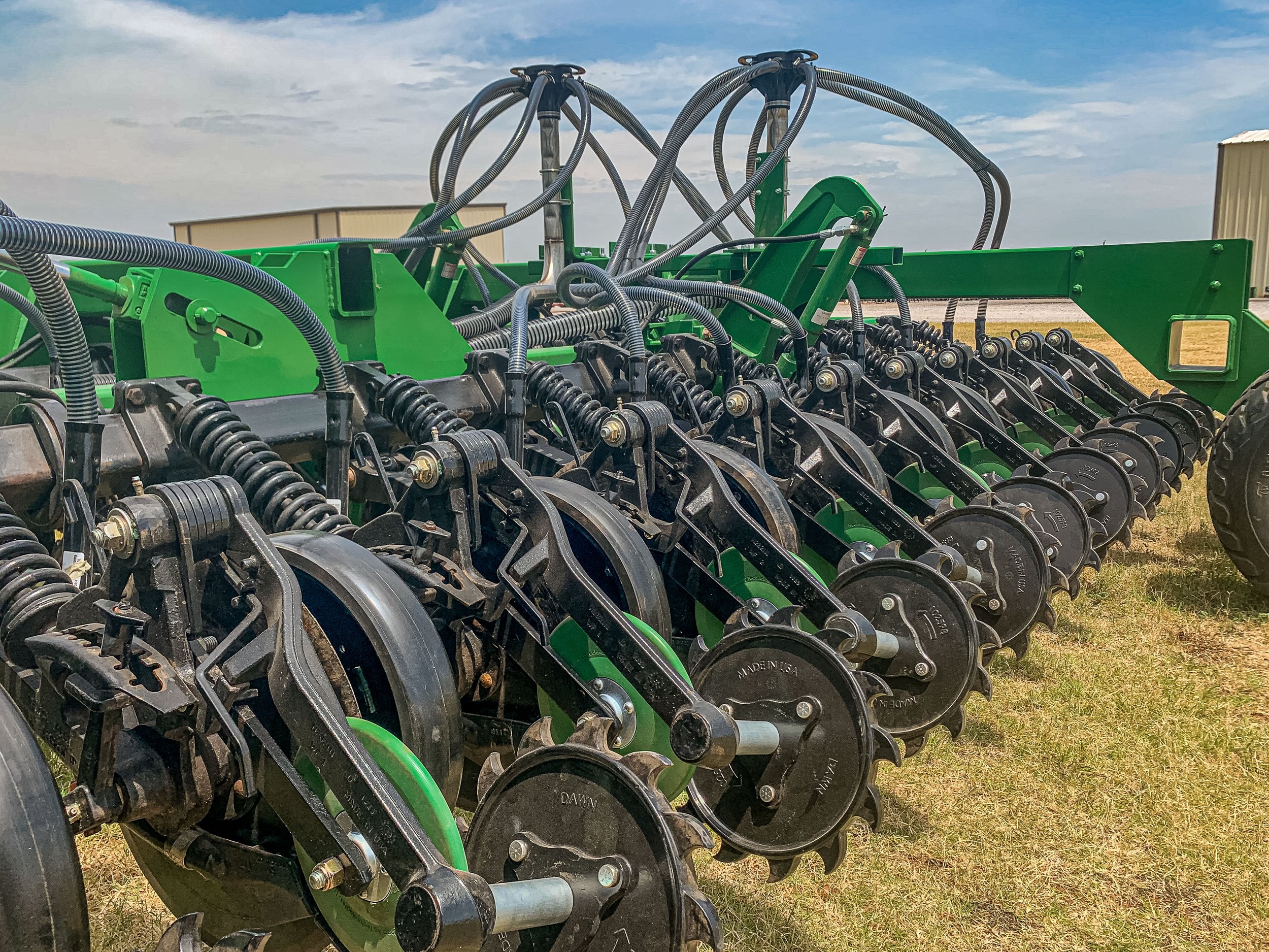A large green agricultural planter machine with multiple black discs and hydraulic hoses, situated outdoors on a grassy field under a partly cloudy sky.