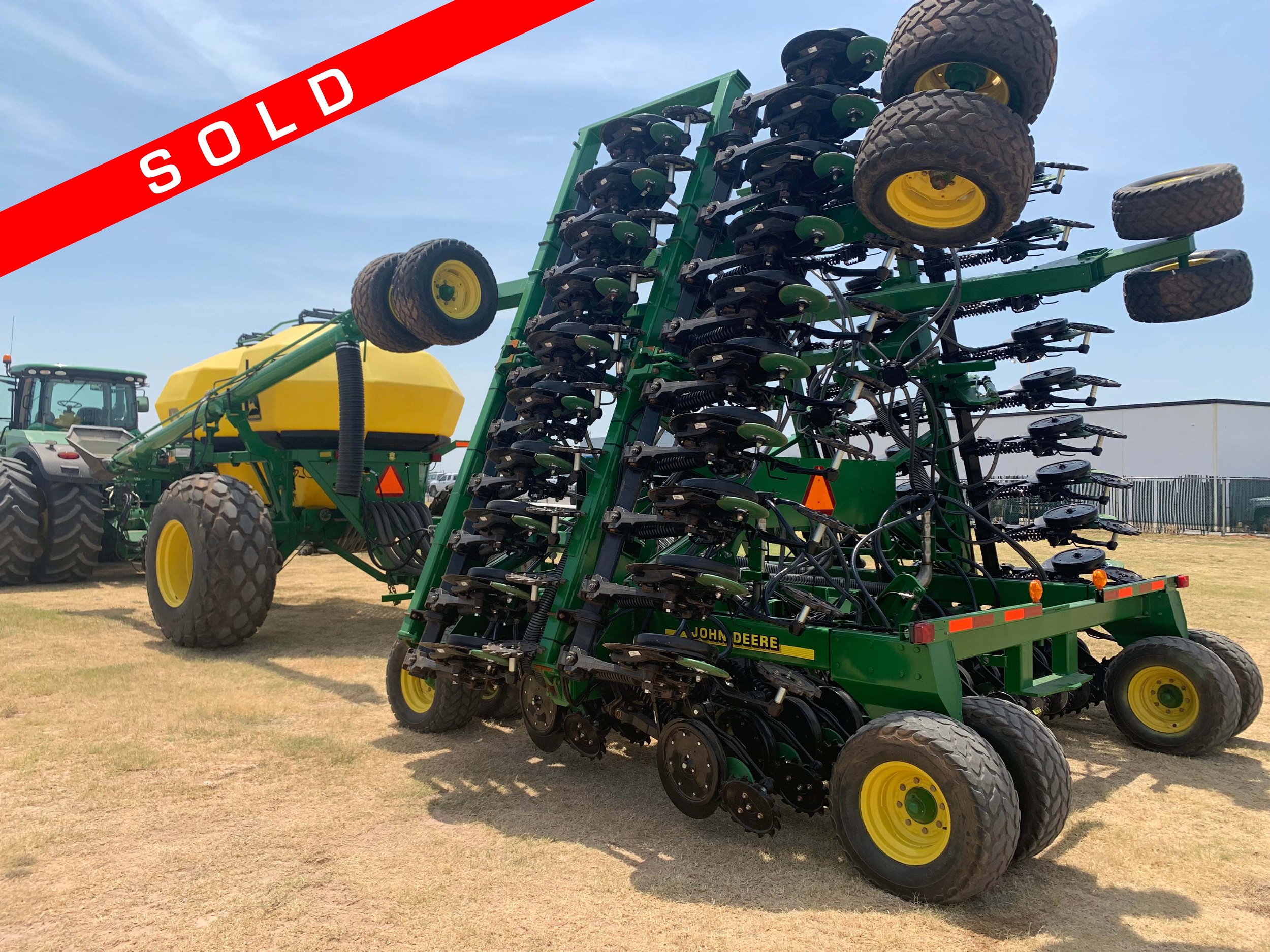 Green and yellow John Deere agricultural sprayer with multiple spray booms, parked on a grassy field with a large tractor in the background, and a red 'SOLD' banner across the top.