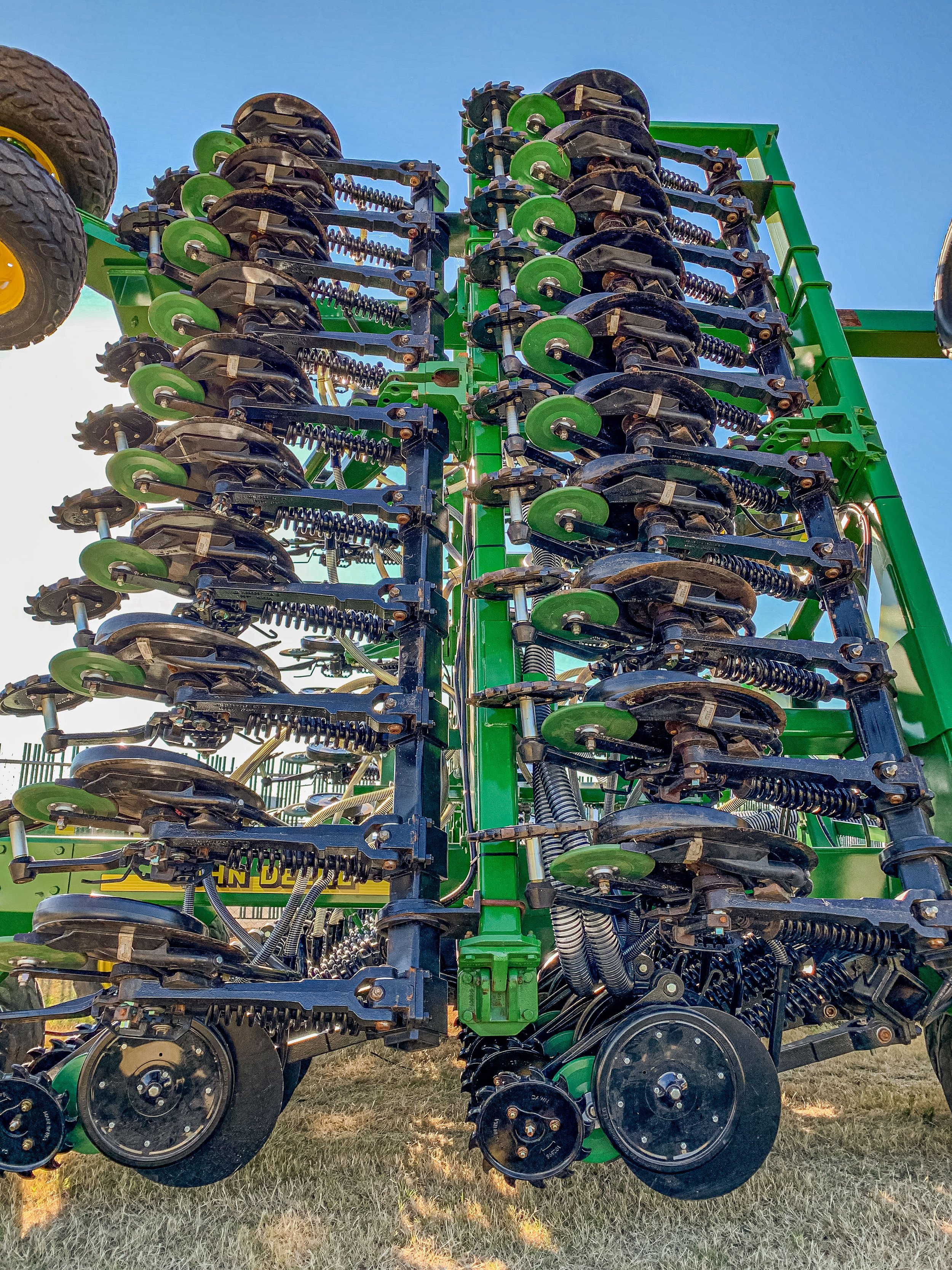 Close-up of a large green and black agricultural planter with multiple disc openers and springs, parked on grass under a blue sky.