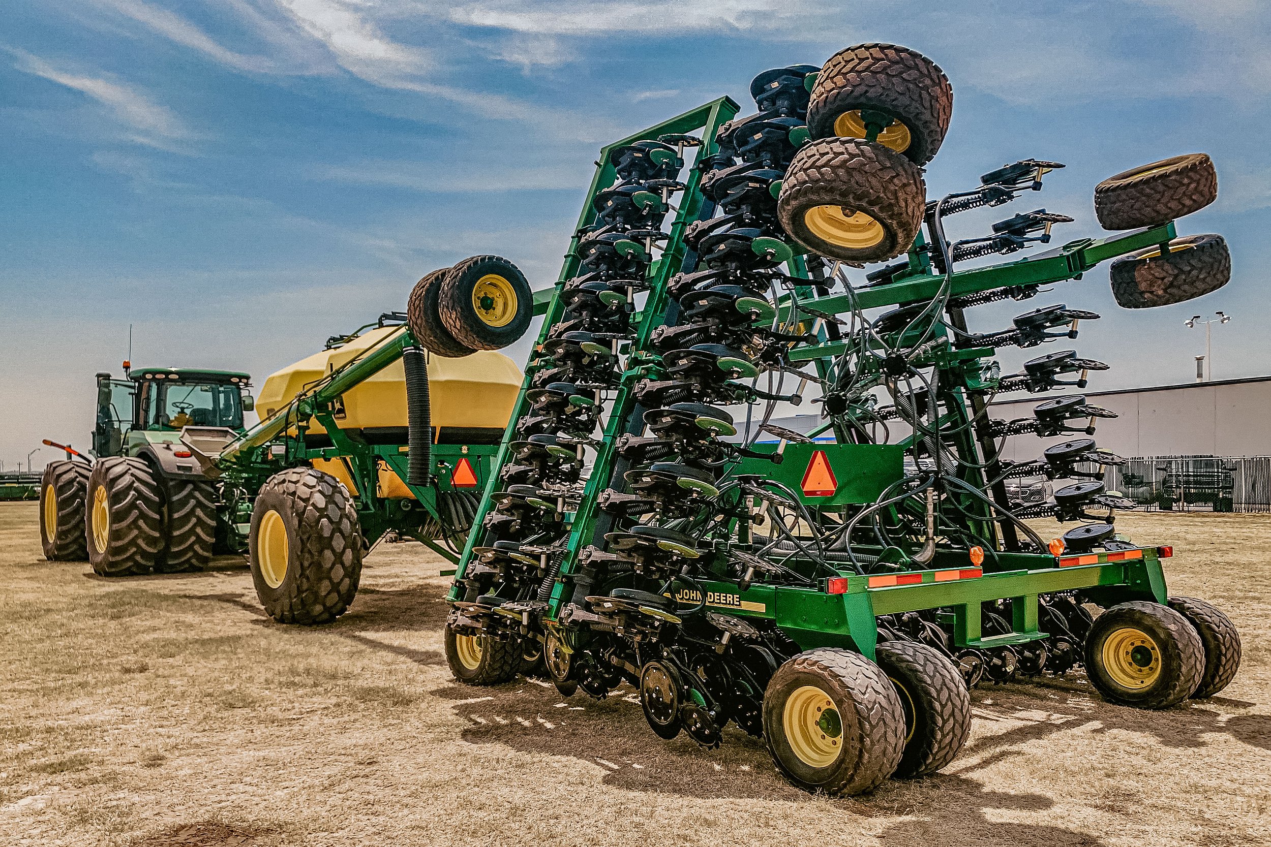 A large green and yellow John Deere agricultural tractor attached to a planting or soil preparation implement on a farmland with a clear blue sky.