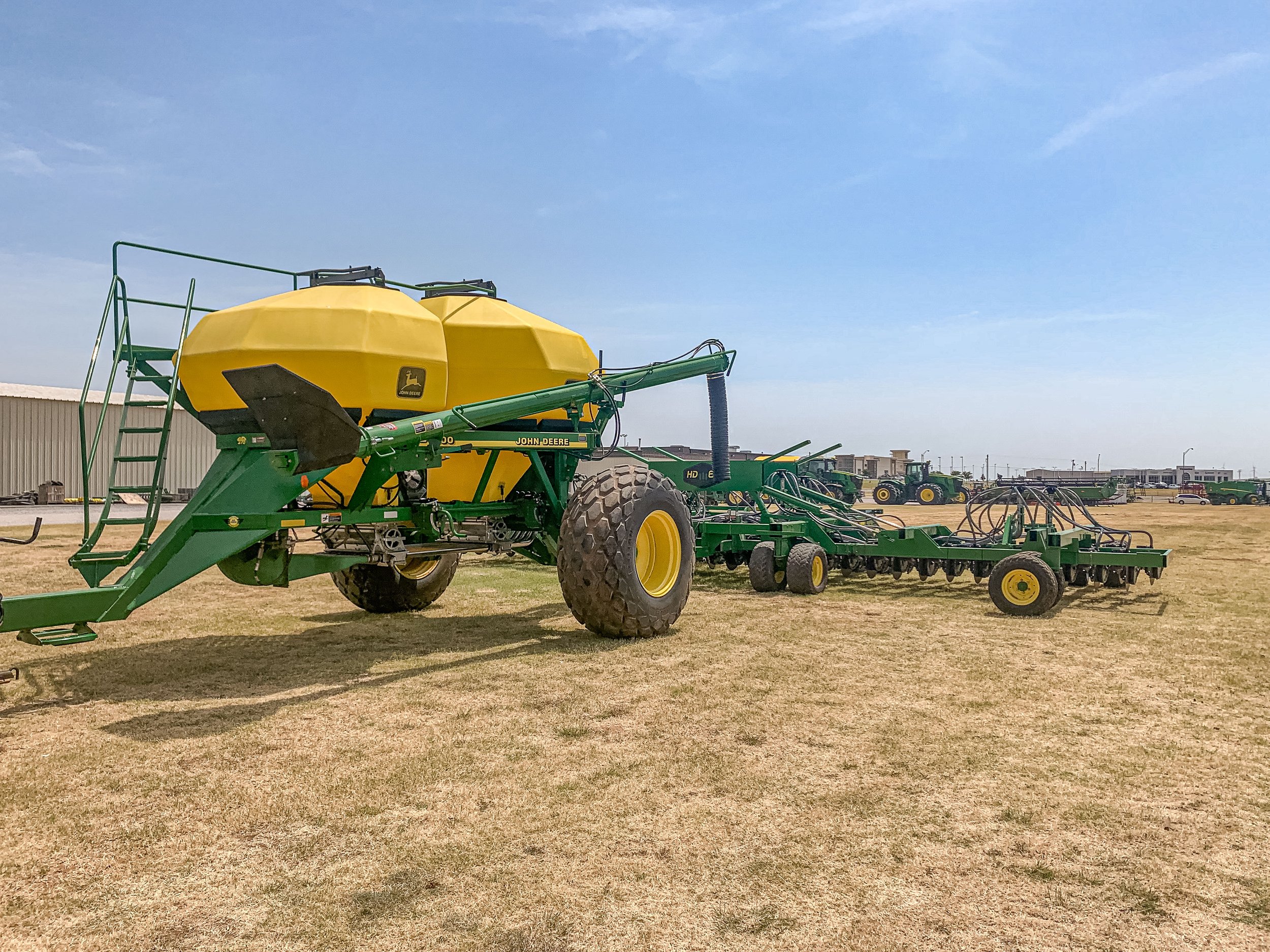 Agricultural equipment including a large green John Deere air seeder with yellow tanks and other farm machinery in the background on a dry field.