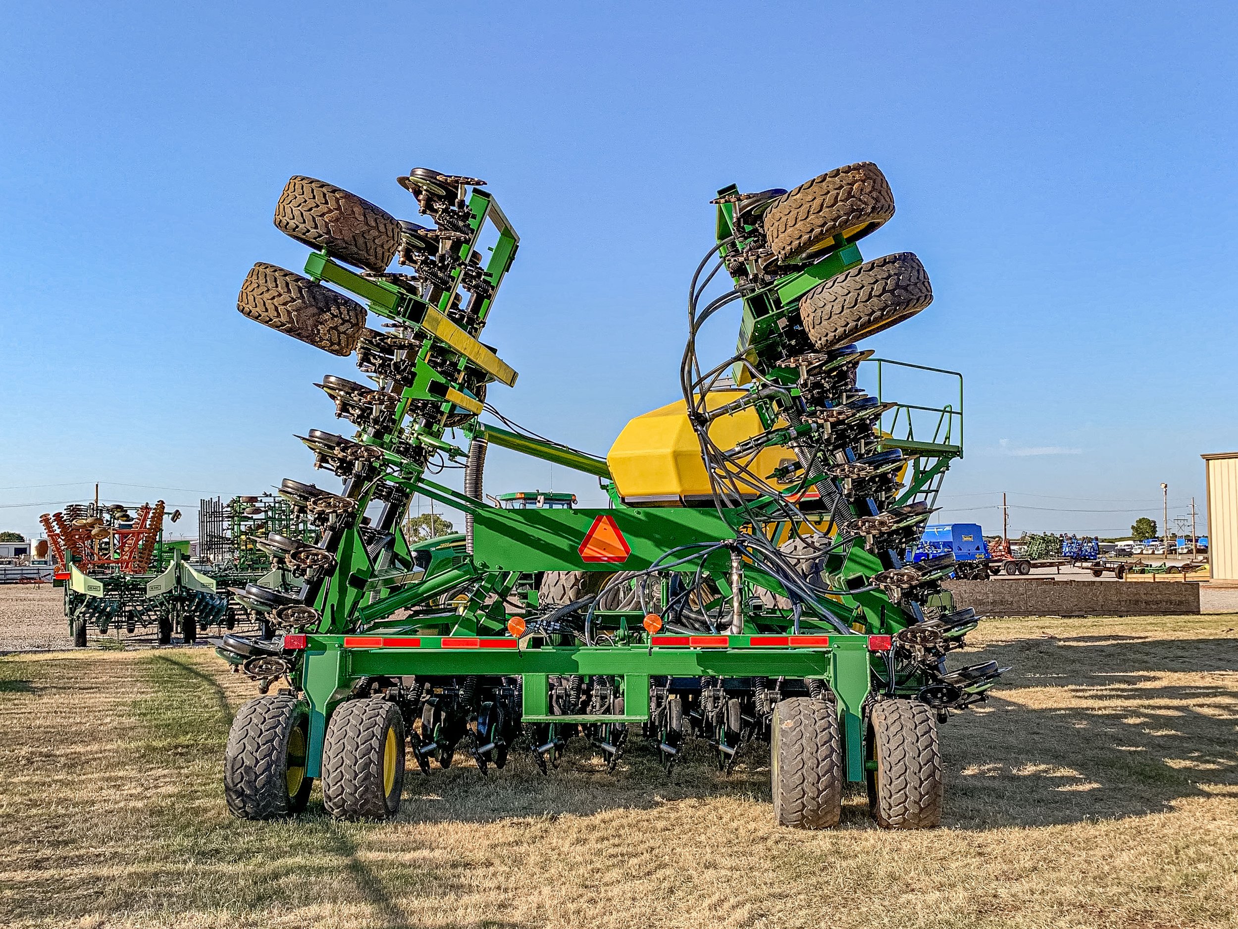 A large green and yellow farm implement with multiple tires and hydraulic hoses on a grassy and gravel lot under a clear blue sky.