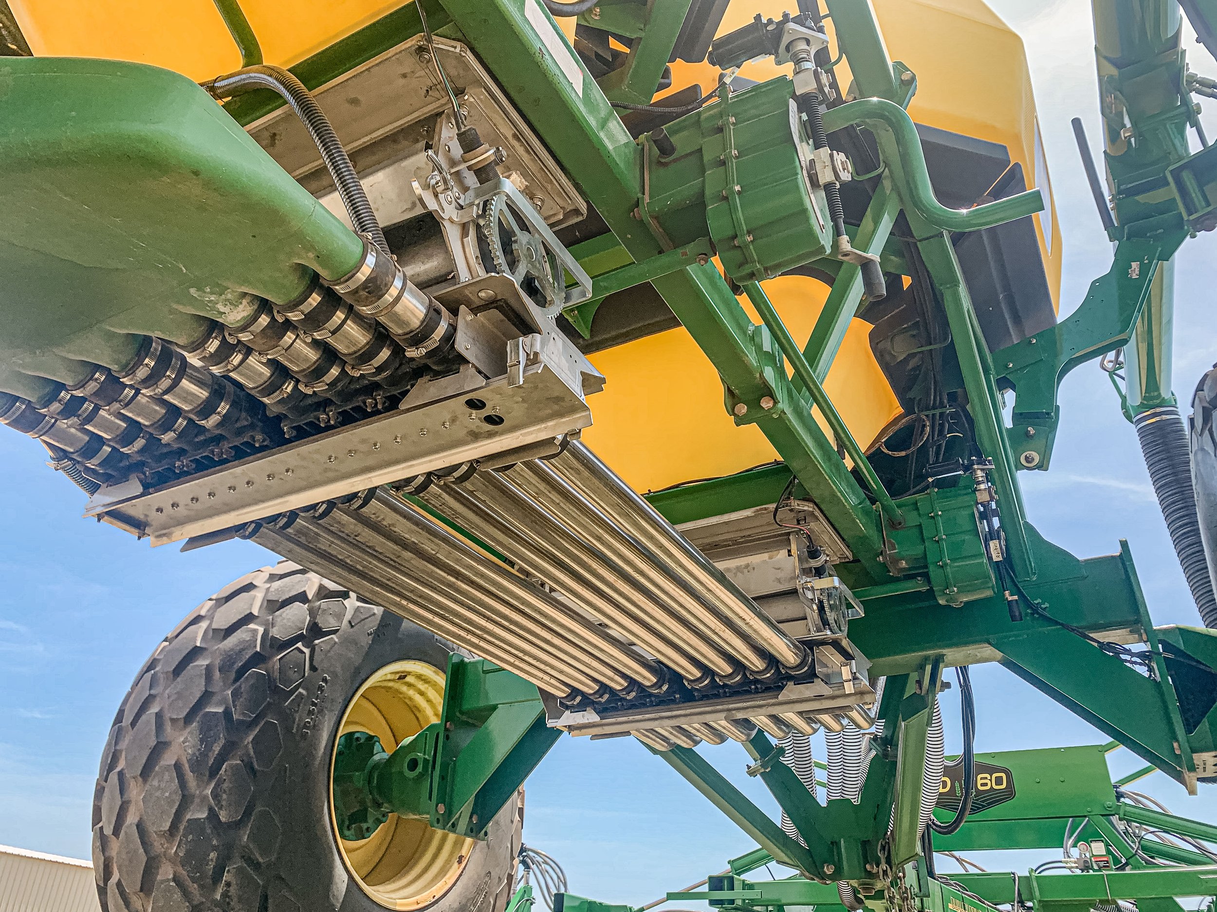 Low-angle view of a large green and yellow agricultural sprayer with metal pipes and mechanical components visible under the spray tank.