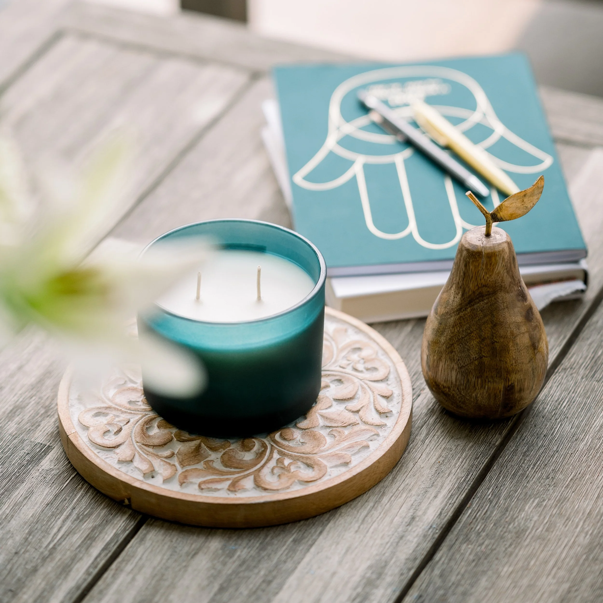 A teal candle in a glass holder on a decorative wooden tray, next to a wooden pear-shaped object with a small metallic leaf, and a blue notebook with white line art of a flower, all arranged on a wooden surface.
