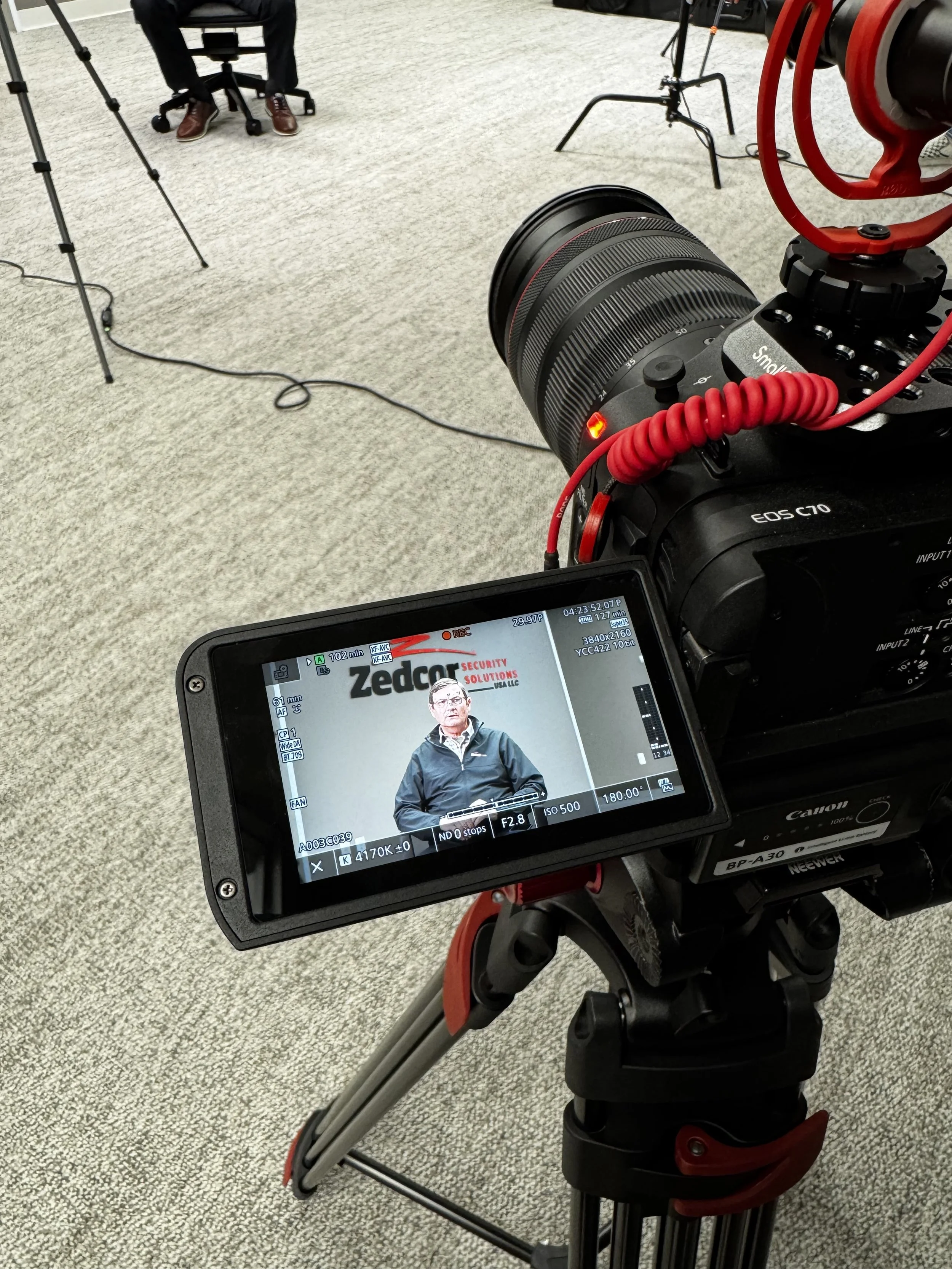 A professional video camera on a tripod filming a man against a backdrop with a monitor displaying his image. The man is wearing a jacket and glasses, standing indoors on a carpeted floor, with equipment and chairs visible in the background.