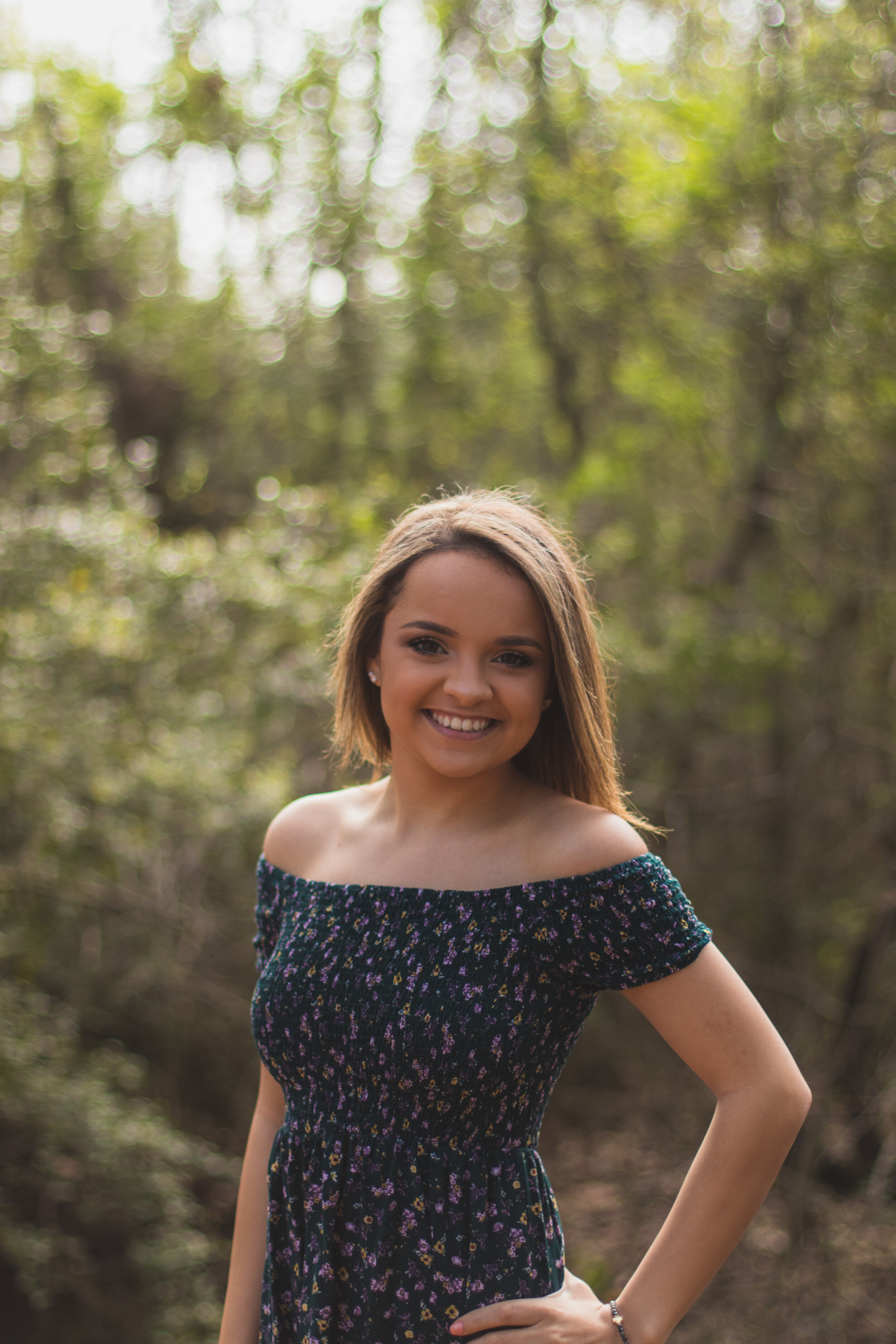 Young woman with shoulder-length light brown hair smiling outdoors in a forest, wearing an off-the-shoulder floral dress.